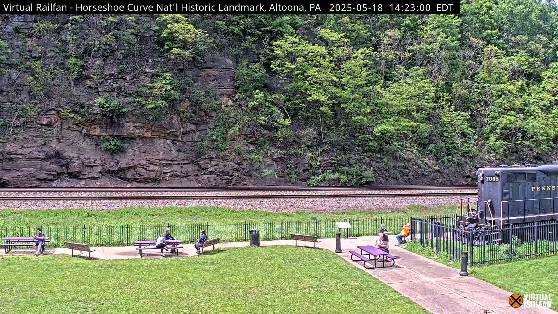 A view of a grassy area with picnic tables and people, situated beside railroad tracks and a rocky hillside with lush green vegetation, under a sunny sky.
