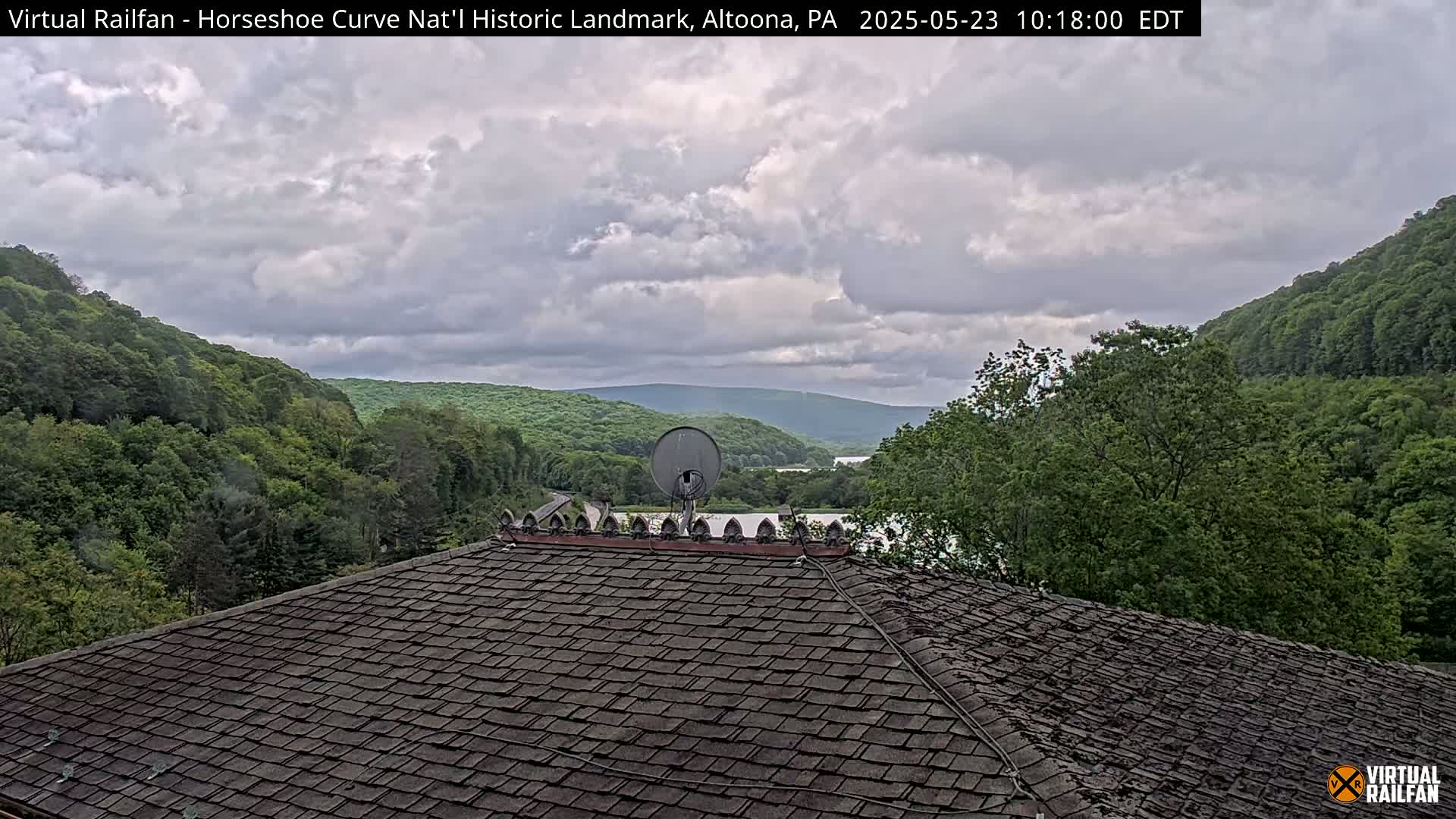 A dark gray shingled roof with a satellite dish sits atop a building overlooking a valley of lush green trees and a body of water under a cloudy sky.