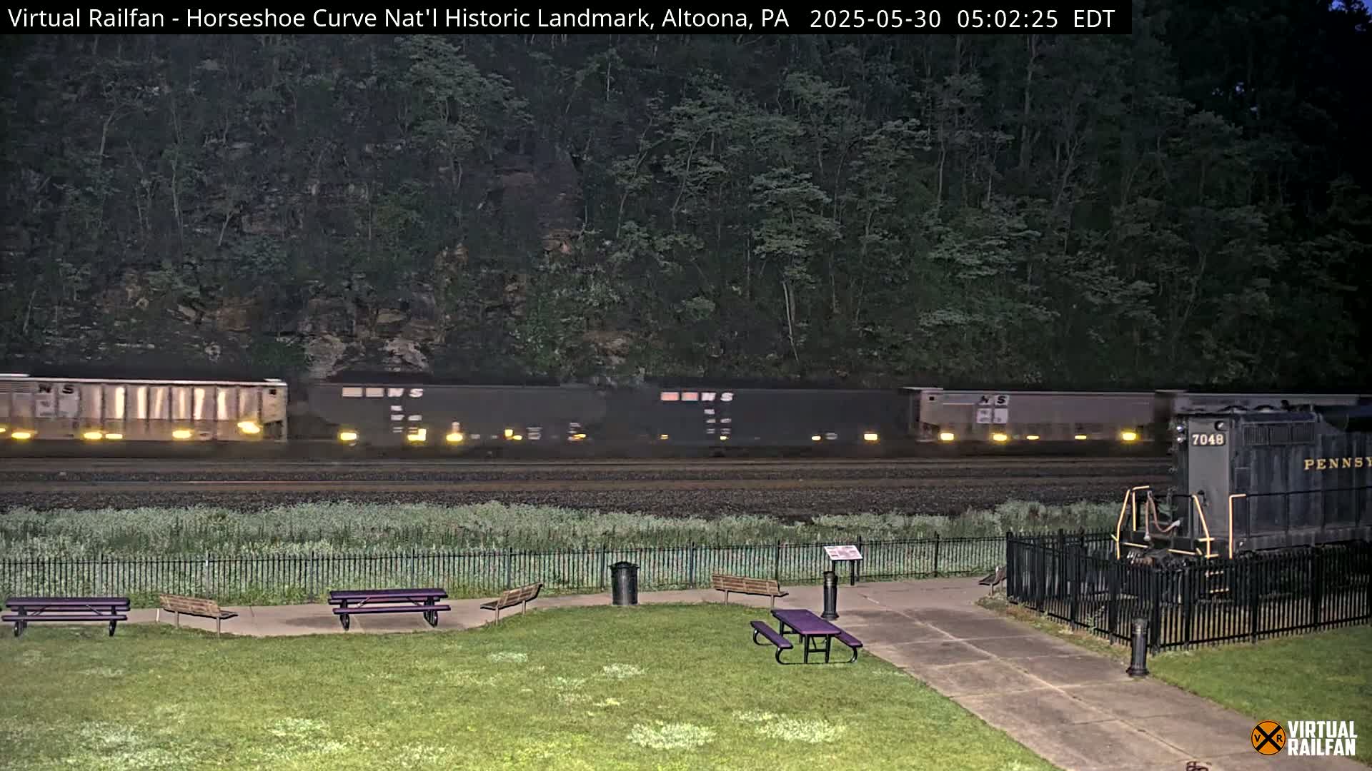 A long freight train passes by a grassy area with picnic tables and a fenced-in area containing a vintage locomotive, all under a dark, pre-dawn sky.