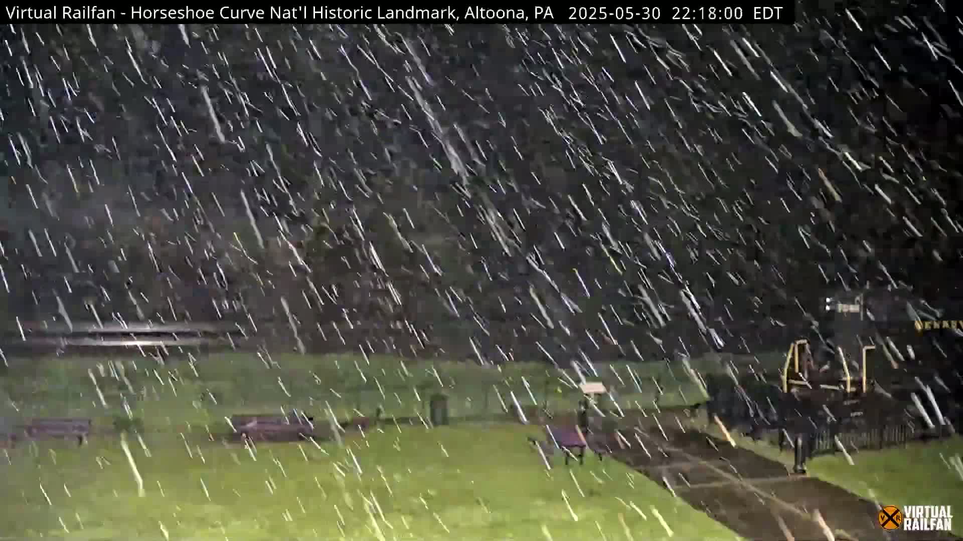 A nighttime scene of a grassy area with park benches and a train in the background, heavily covered in falling snow.