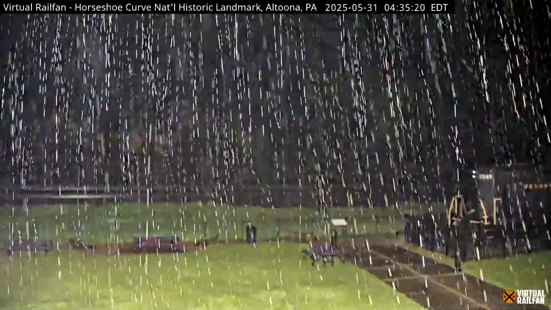 A dark, nighttime scene of a grassy area with park benches and a partially visible train is shown under heavy rainfall.