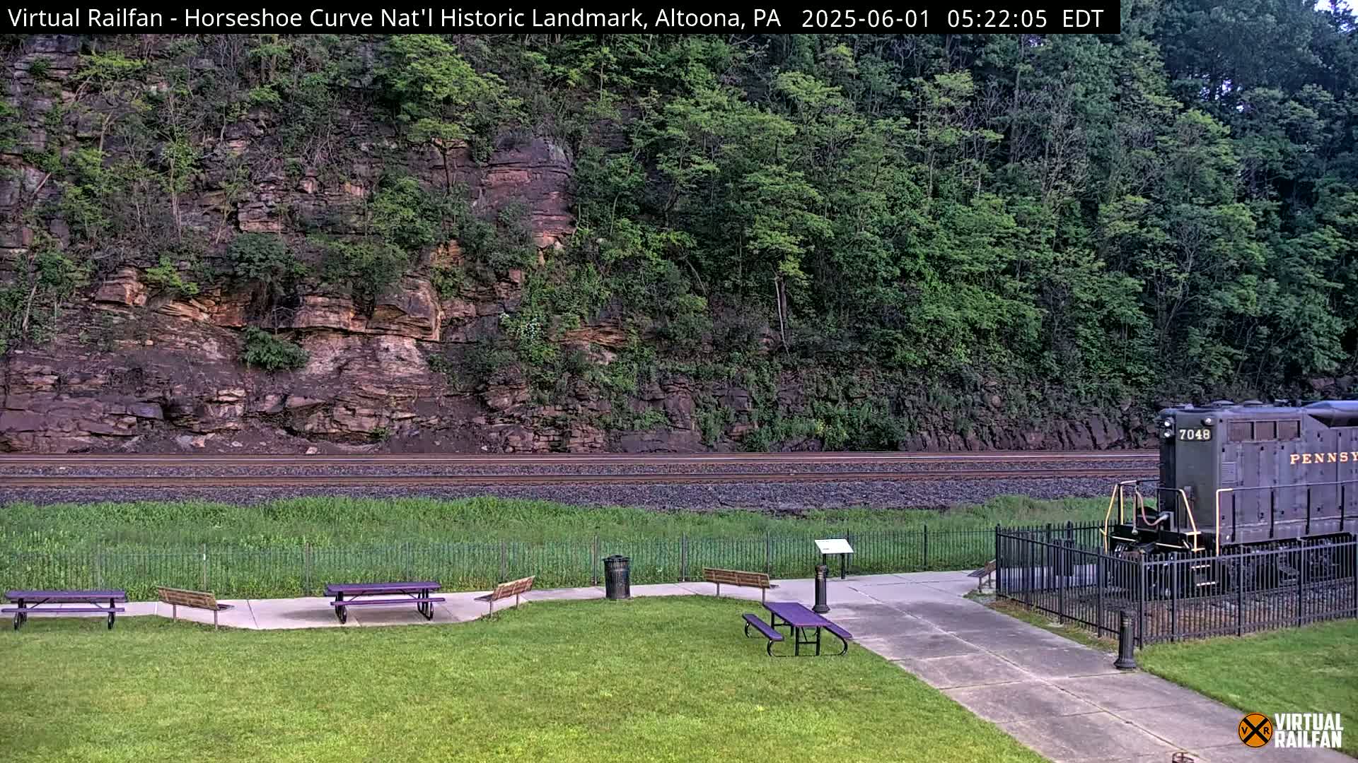 A train sits on railway tracks in front of a grassy area with picnic tables, all set against a backdrop of a rocky hillside covered in lush green trees under a partly cloudy sky.