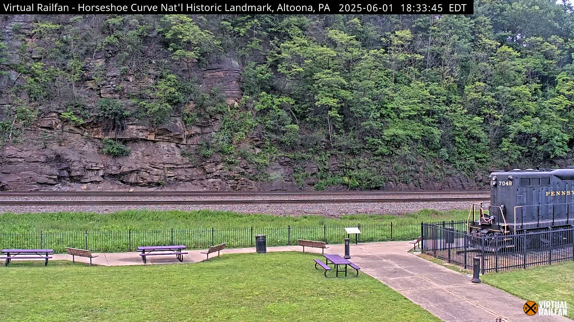 A dark-colored train sits on railway tracks beside a grassy area with picnic tables, all situated before a rocky hillside with lush green vegetation under an overcast sky.