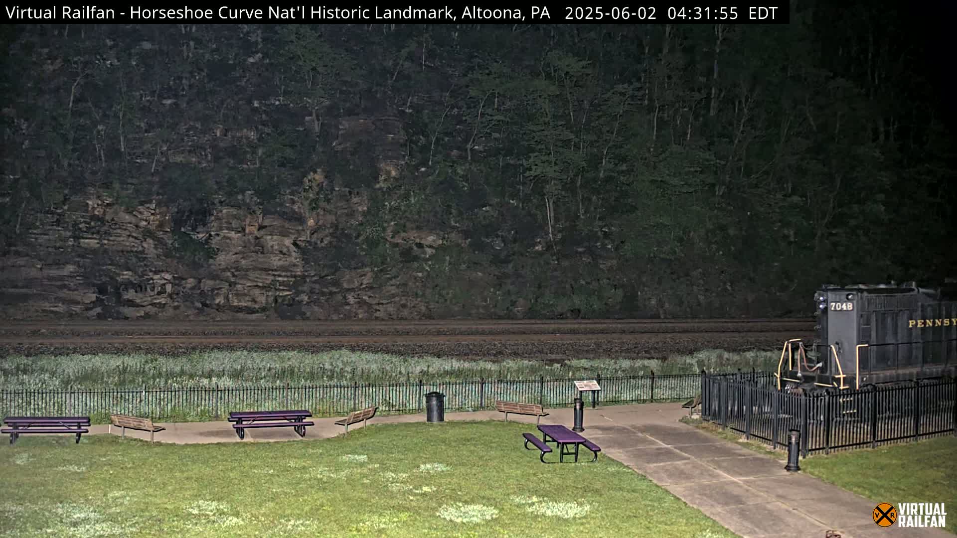 A nighttime view of a grassy area with picnic tables, a fenced-in train engine, and a railroad track leading to a rocky hillside covered in trees.