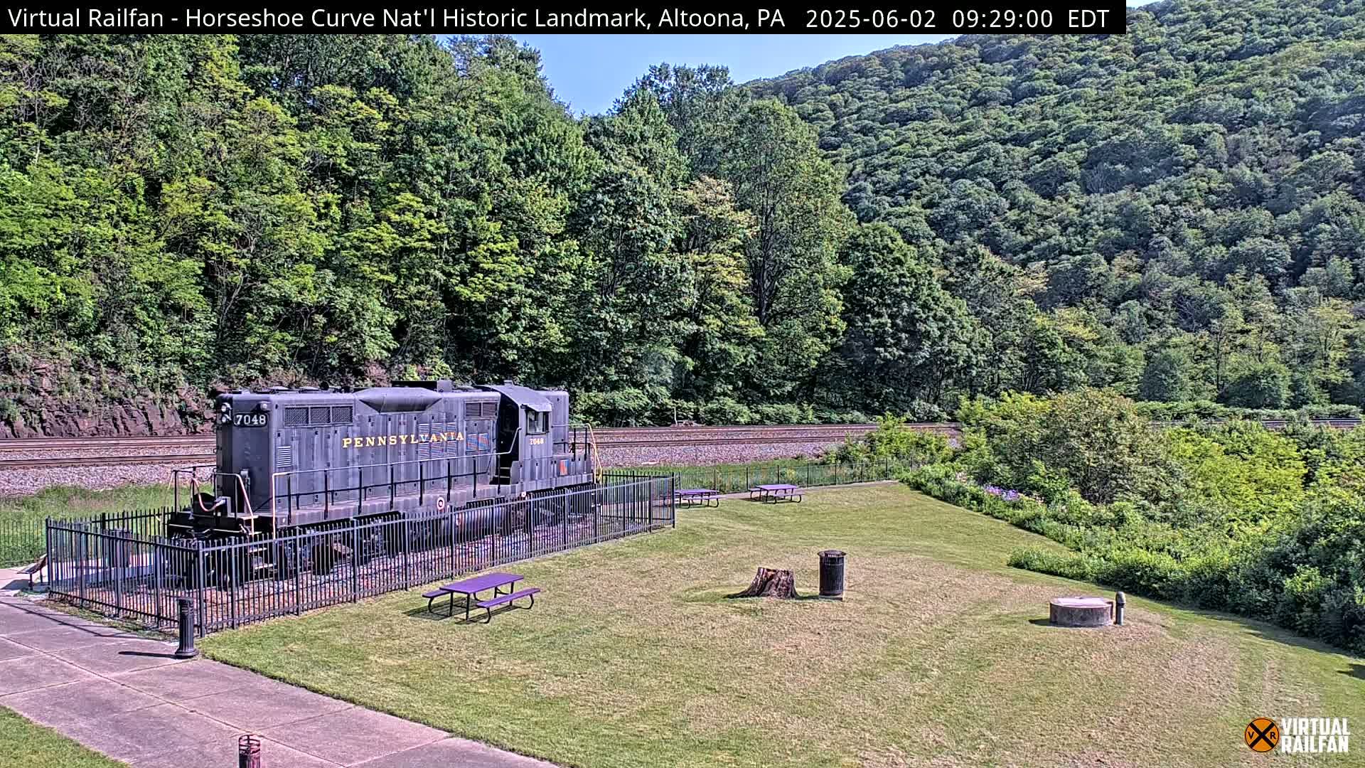 A Pennsylvania Railroad diesel locomotive sits behind a black fence on a grassy area, next to a picnic area, with a lush green hillside in the background on a sunny day.