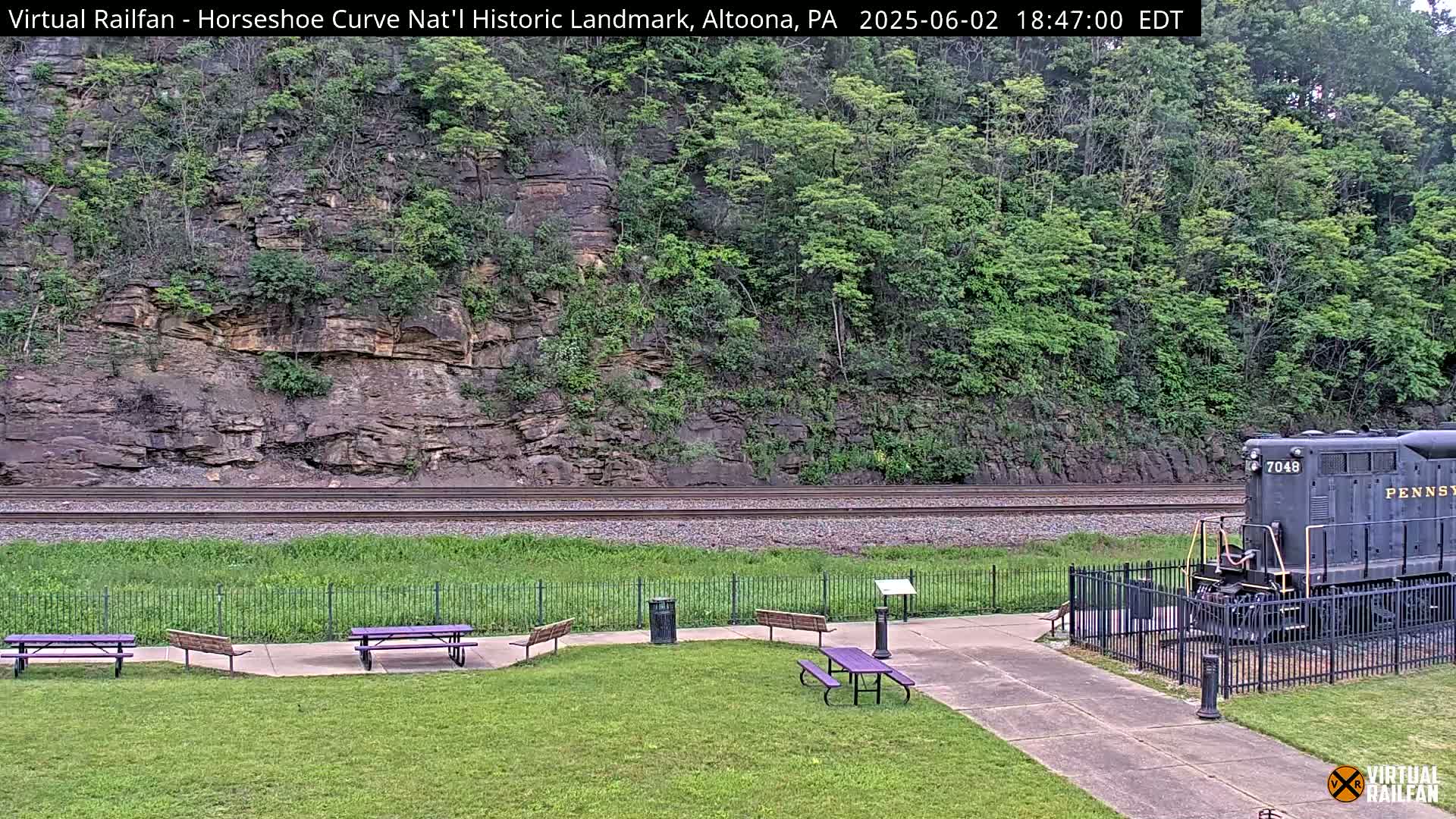 A grassy area with picnic tables and a black diesel locomotive sits beside railroad tracks that curve around a rocky hillside covered in lush green trees under a partly cloudy sky.