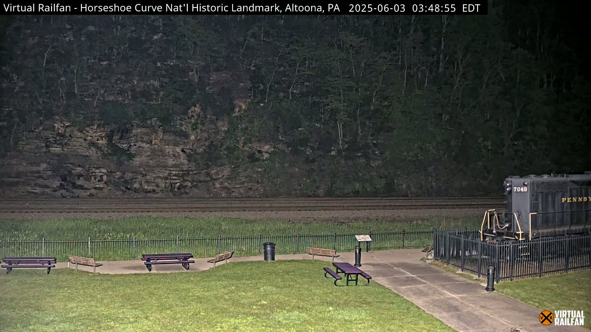 A grassy area with picnic tables overlooks a railway track where a train is parked at night, beyond which is a steep, wooded hillside.