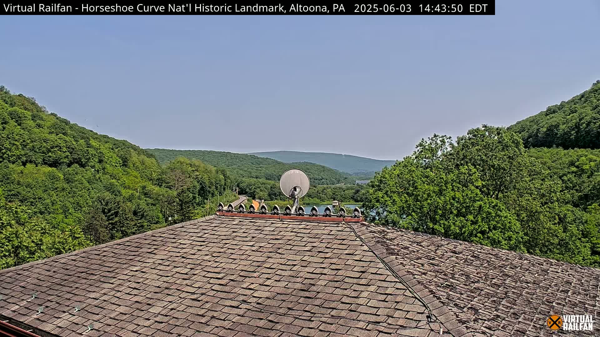 A brown shingled roof with a satellite dish overlooks a valley of lush green trees under a clear blue sky.