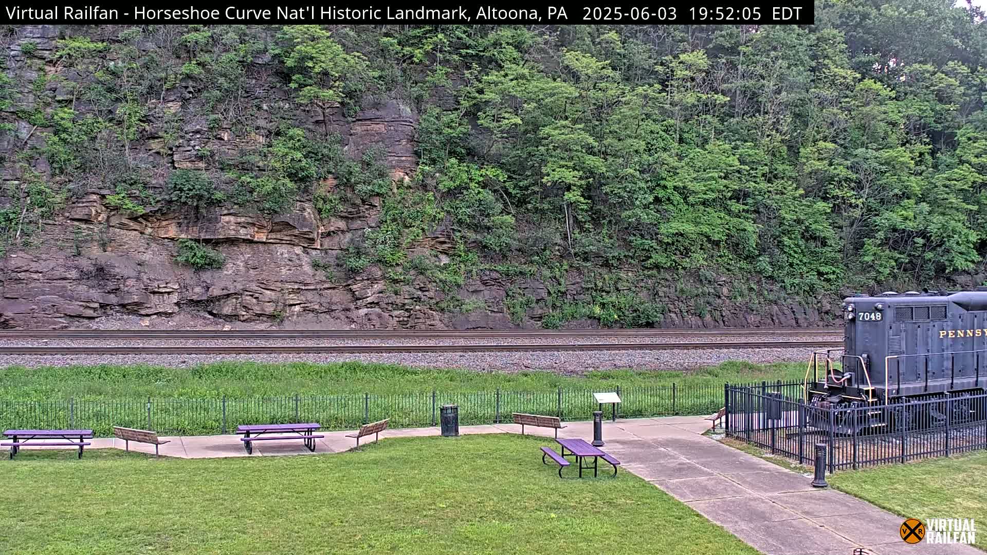 A dark-colored locomotive sits near a grassy area with picnic tables beside railroad tracks that curve along a steep, rocky hillside covered in green vegetation under a partly cloudy sky.