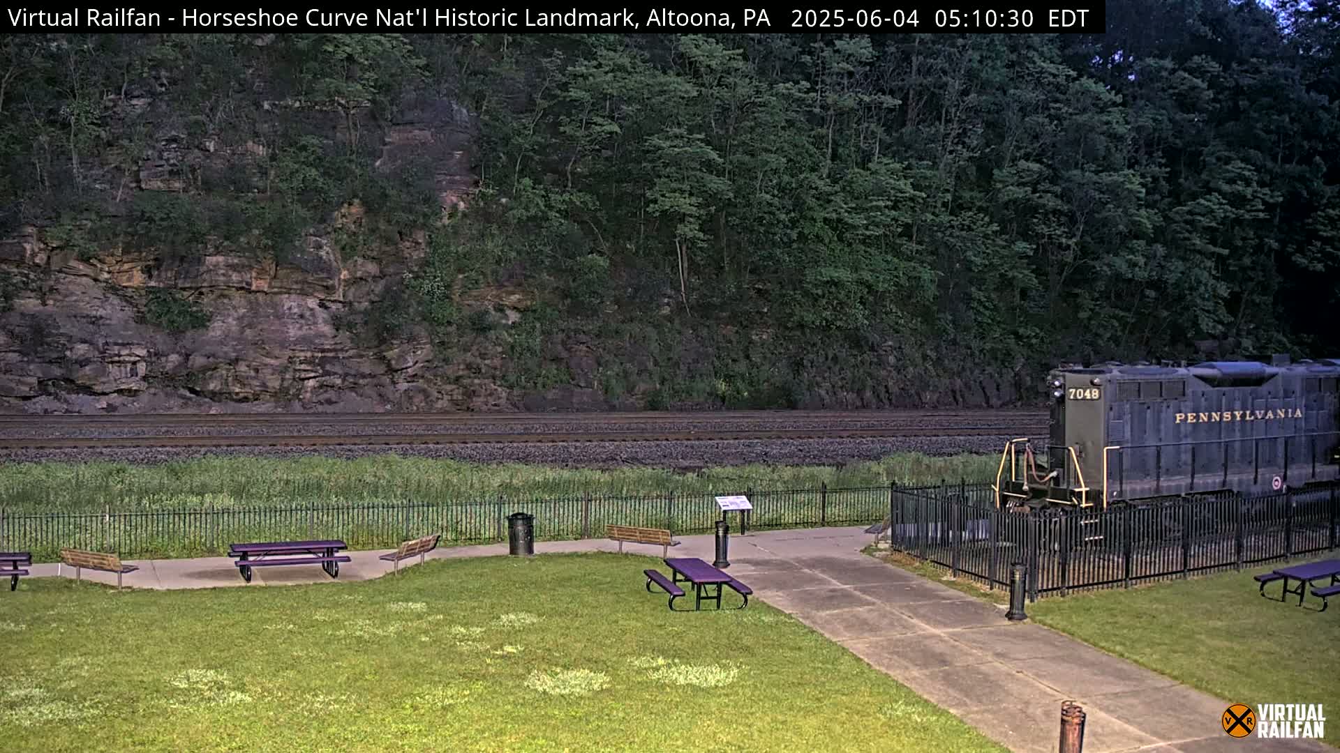 A dark-green Pennsylvania Railroad locomotive sits behind a black fence in a park-like setting next to a grassy area with picnic tables, all in front of a rocky hillside under a dusky sky.