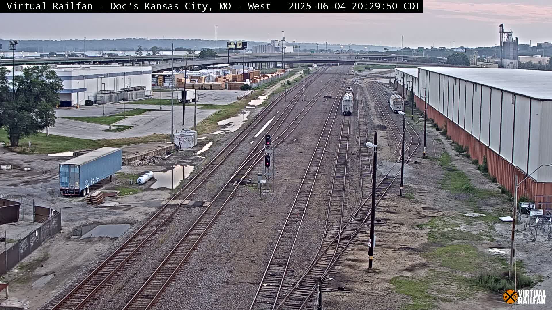 A high-angle, long shot of several railway lines in an industrial area, under a partly cloudy evening sky, shows two trains on separate lines and several puddles on the ground.