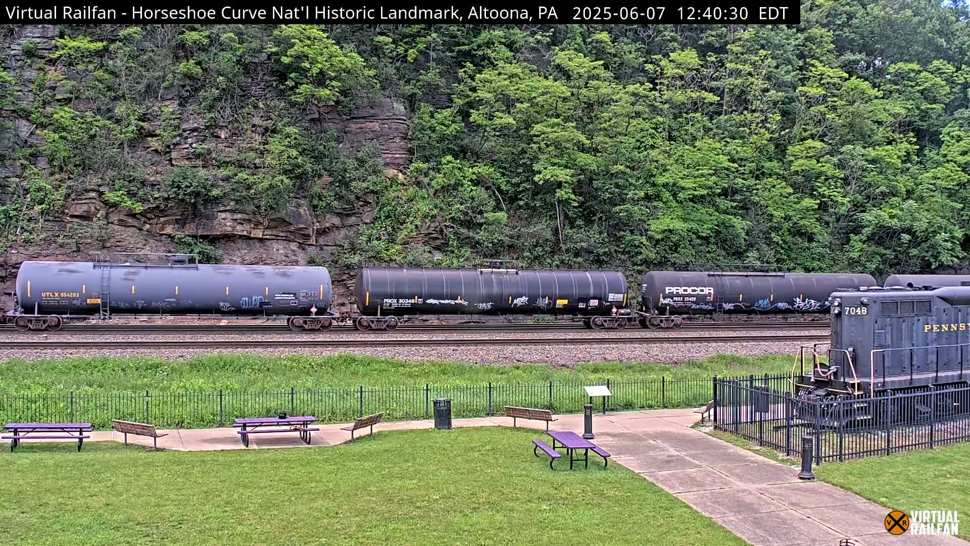 A freight train, consisting of several tanker cars and a locomotive, is traveling along a railway track, set against a backdrop of a rocky hillside covered in lush green vegetation.