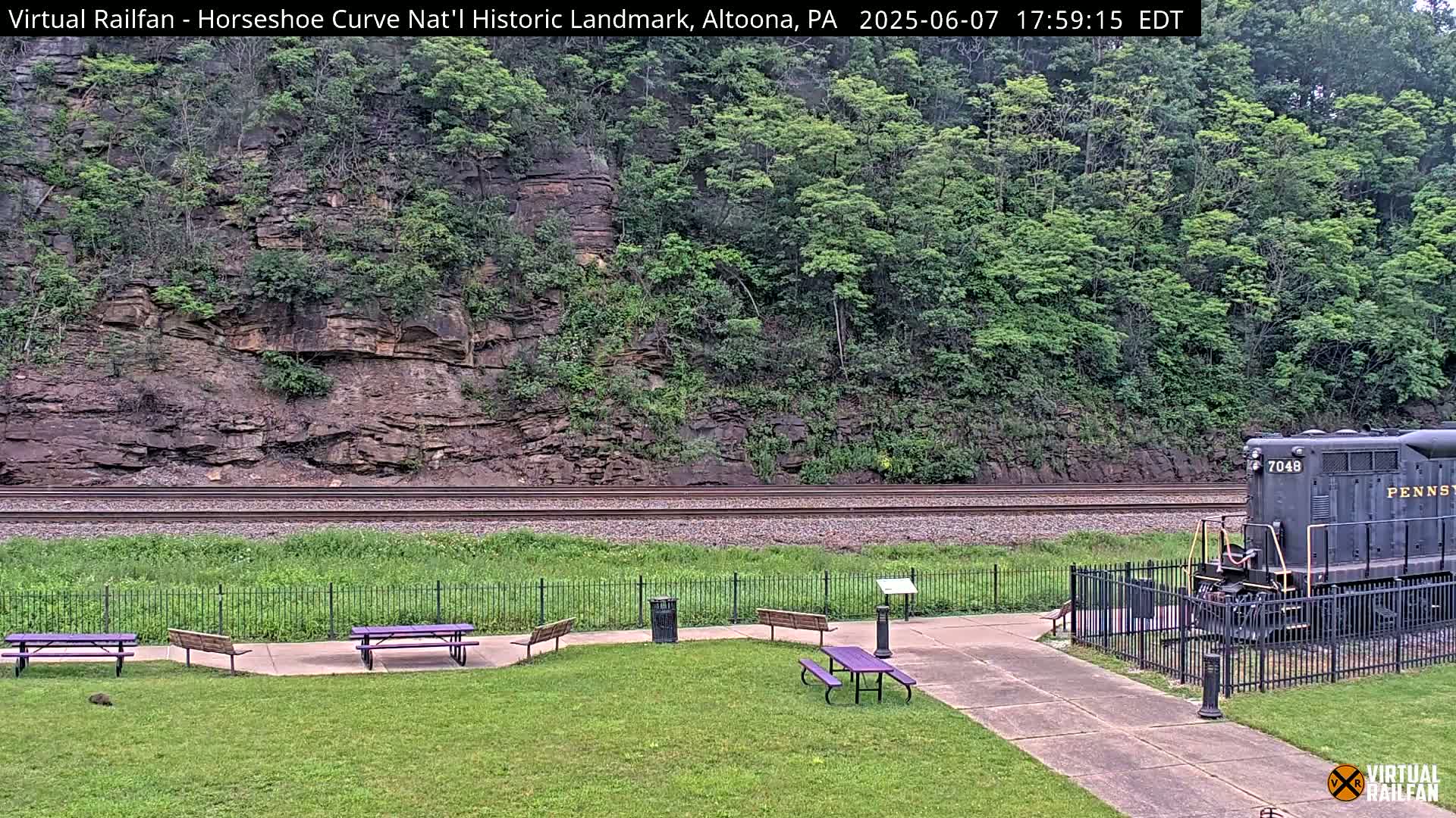A train sits on railroad tracks beside a grassy area with picnic tables, all set against a backdrop of a rocky hillside covered in green foliage.
