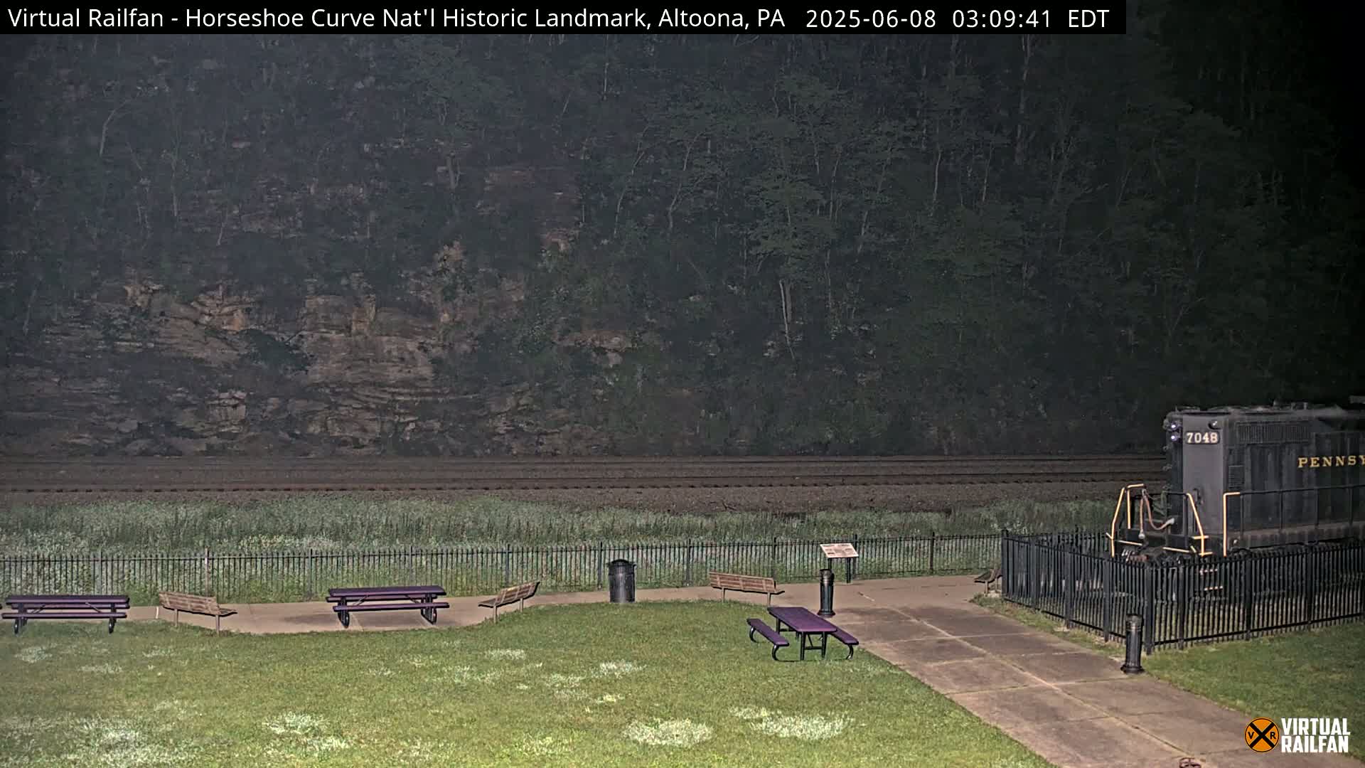 A nighttime view of a grassy area with picnic tables, a trash can, and a black locomotive behind a fence, all set against a backdrop of a wooded hillside and railway tracks.