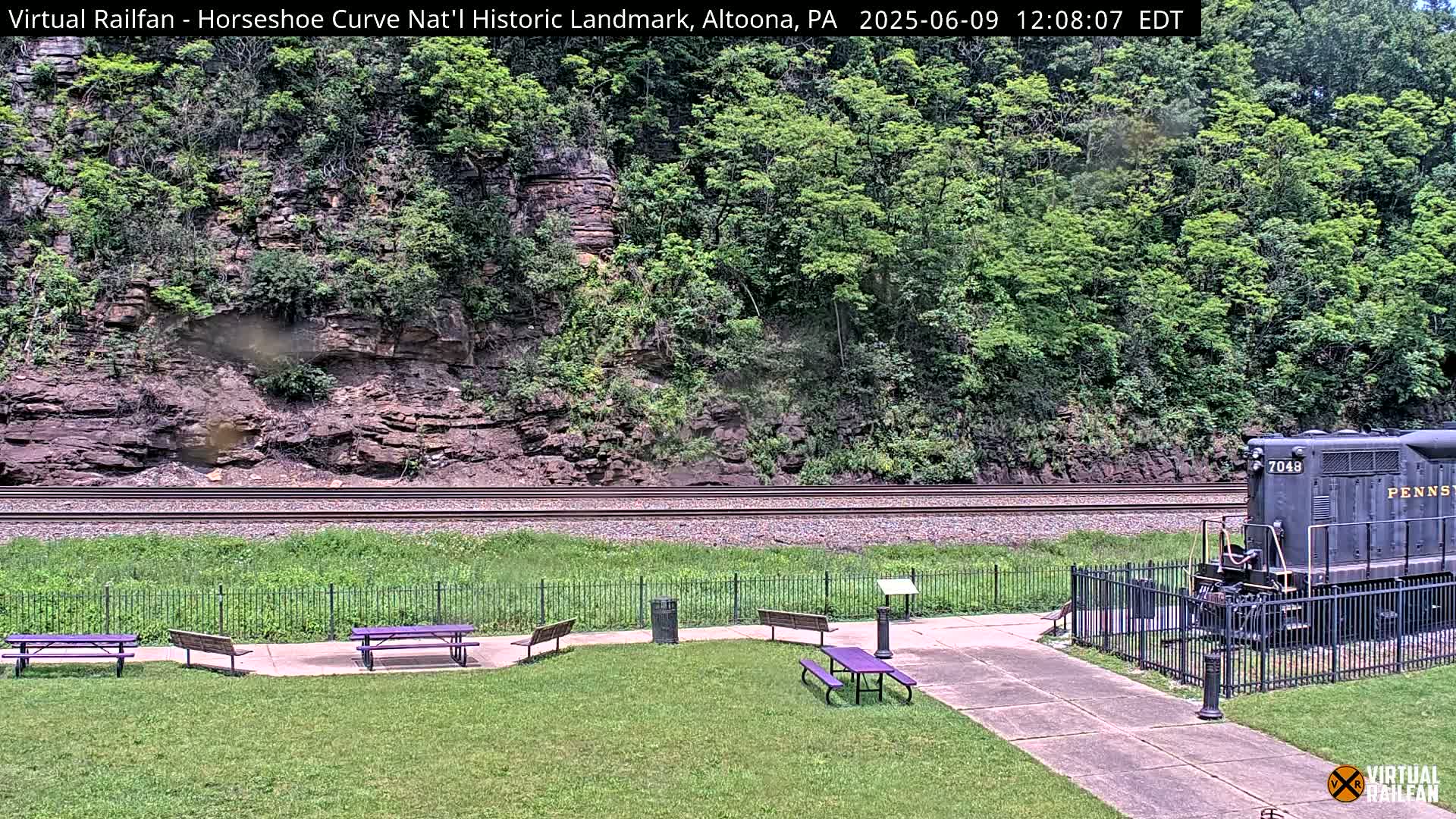 A black diesel locomotive sits near a grassy park area with picnic tables, beside a railway track running along a rocky hillside covered in lush green vegetation.