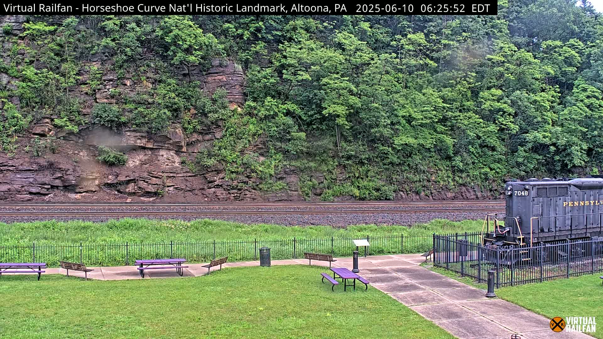 A dark-colored locomotive sits behind a black fence near a grassy area with purple picnic tables, all set against a backdrop of a rocky hillside covered in lush green vegetation.