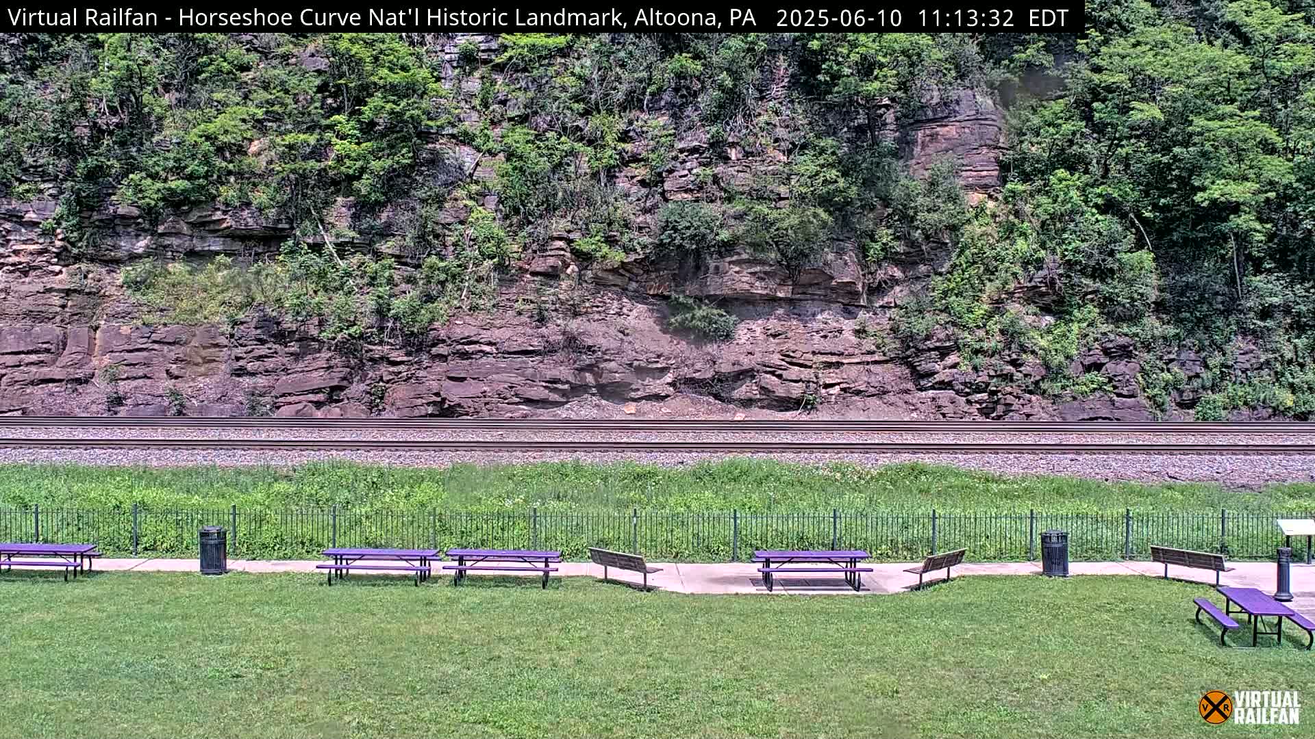 A grassy area with purple picnic tables and a trash can sits before a set of train tracks that run along the base of a rocky hillside covered in green vegetation, under a sunny sky.