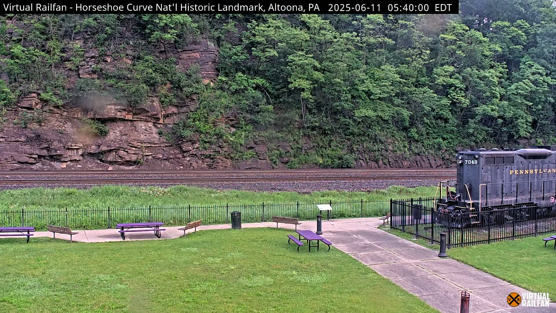 A dark-colored locomotive sits near a grassy area with purple picnic tables in front of a rocky hillside covered in lush green foliage, under overcast skies.