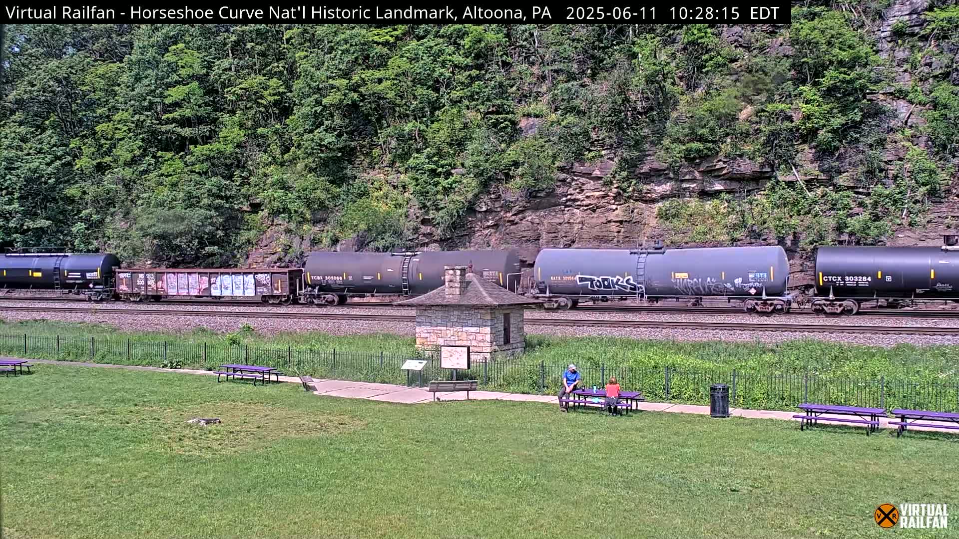 A freight train passes by a stone structure in a grassy park area, backed by a lush, green hillside on a sunny day.