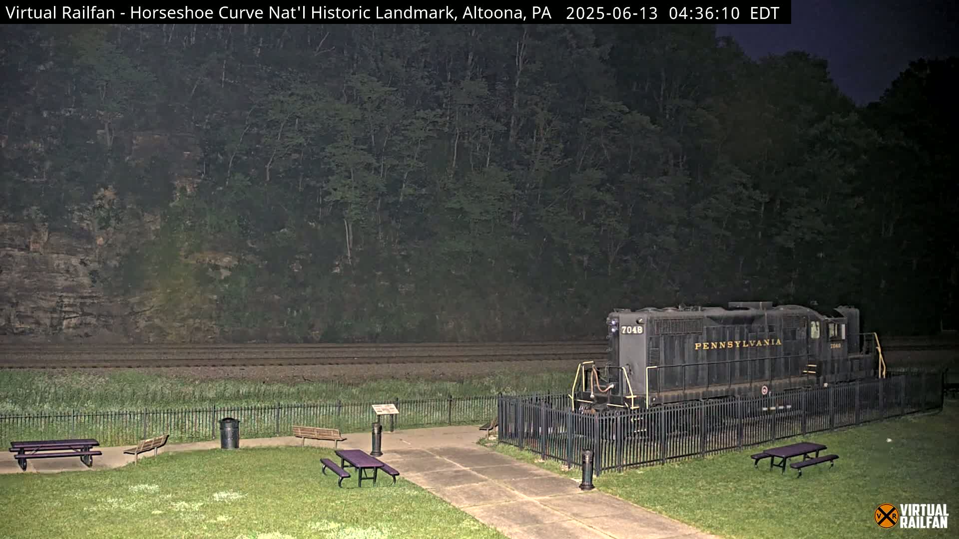 A dark-colored locomotive sits behind a black fence next to a grassy area with picnic tables under a night sky.