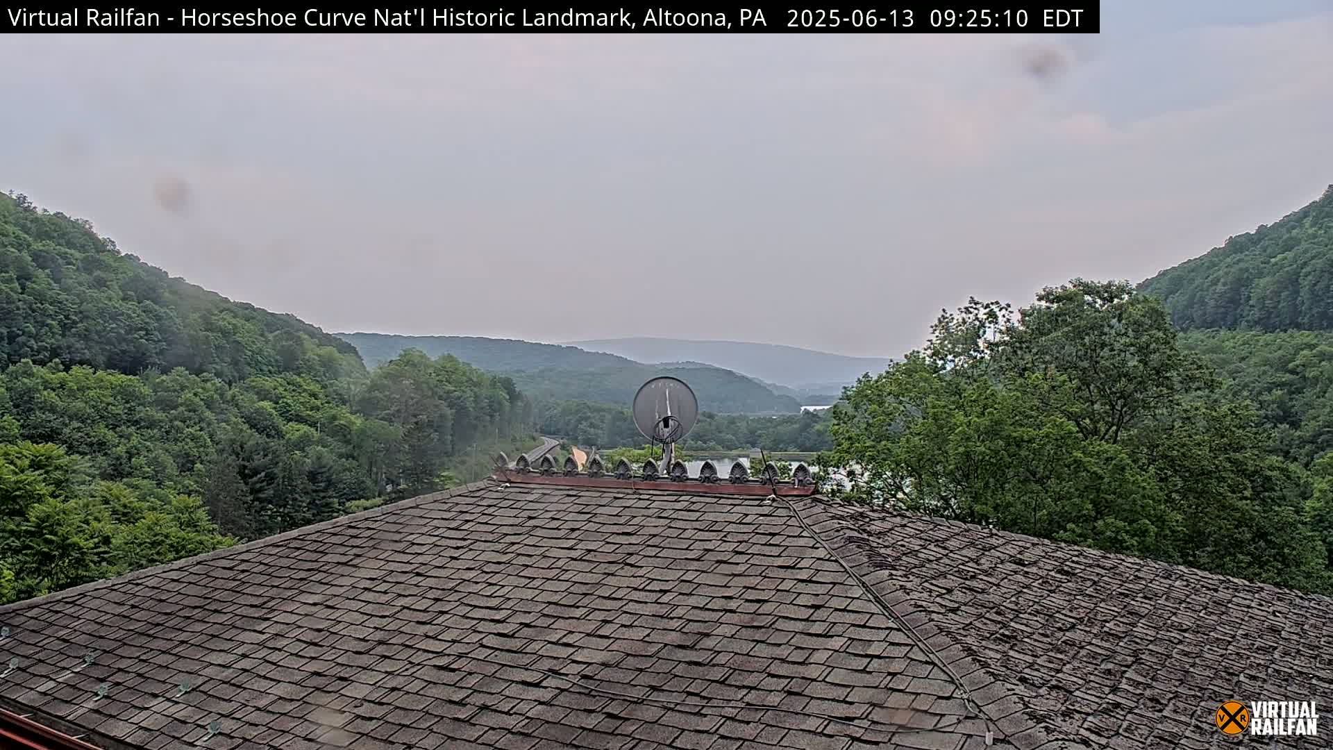 A satellite dish sits atop a shingled roof overlooking a valley of lush green trees under a cloudy sky.
