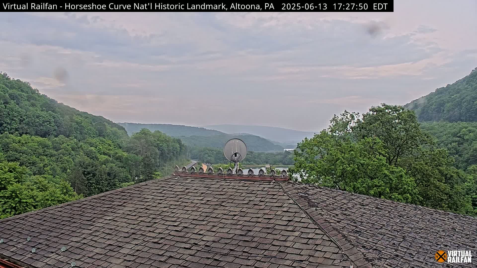 A dark gray shingled roof with a satellite dish sits atop a building overlooking a valley of lush green trees under a mostly cloudy sky.
