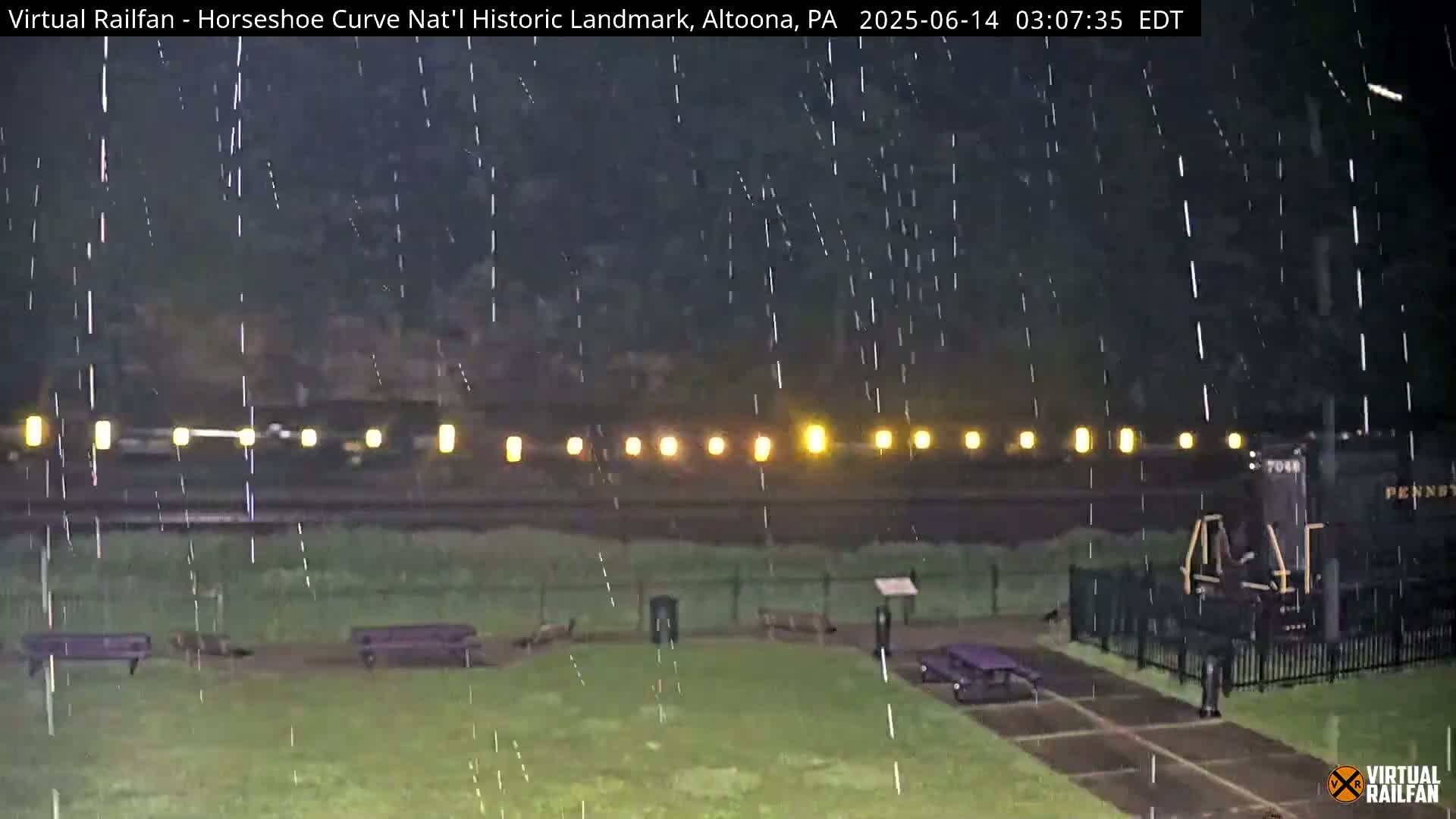 A nighttime view of a train in the rain, with park benches and a fence visible in the foreground.