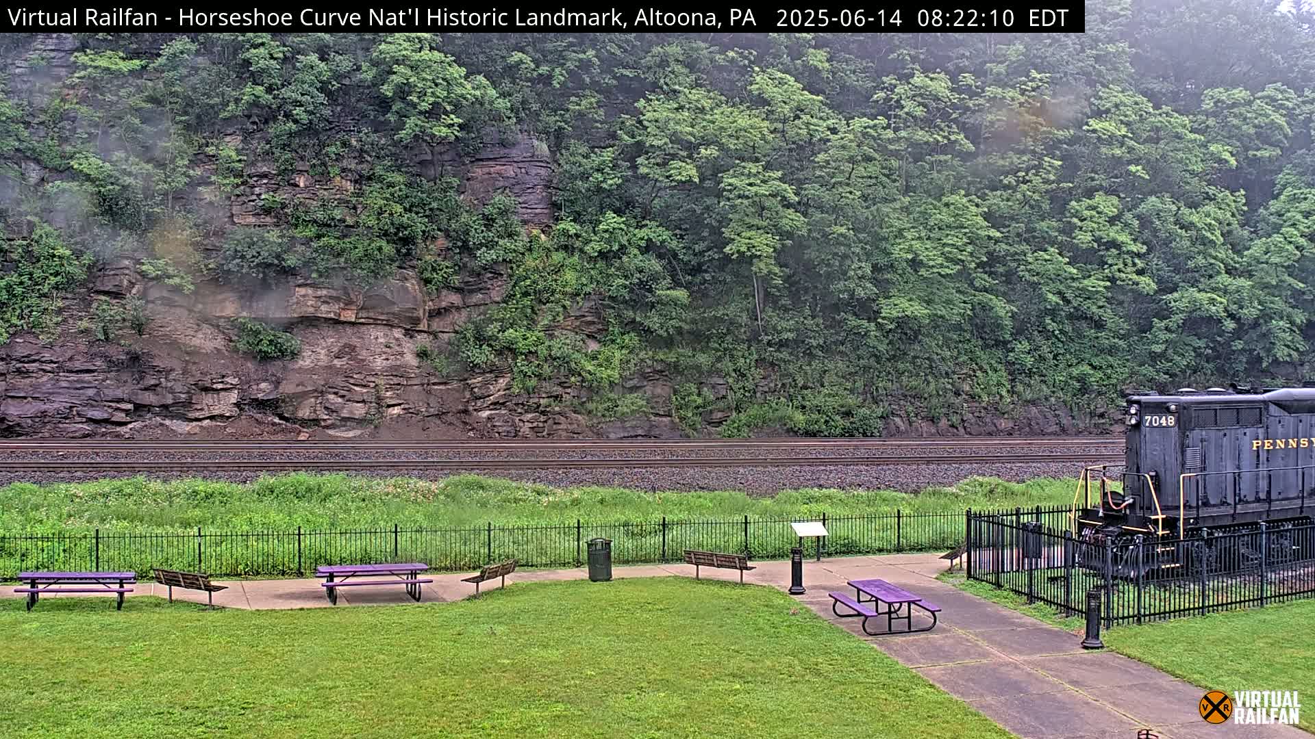 A dark-colored locomotive sits near a grassy area with picnic tables beside railroad tracks that curve alongside a rocky hillside covered in green foliage, under overcast skies.