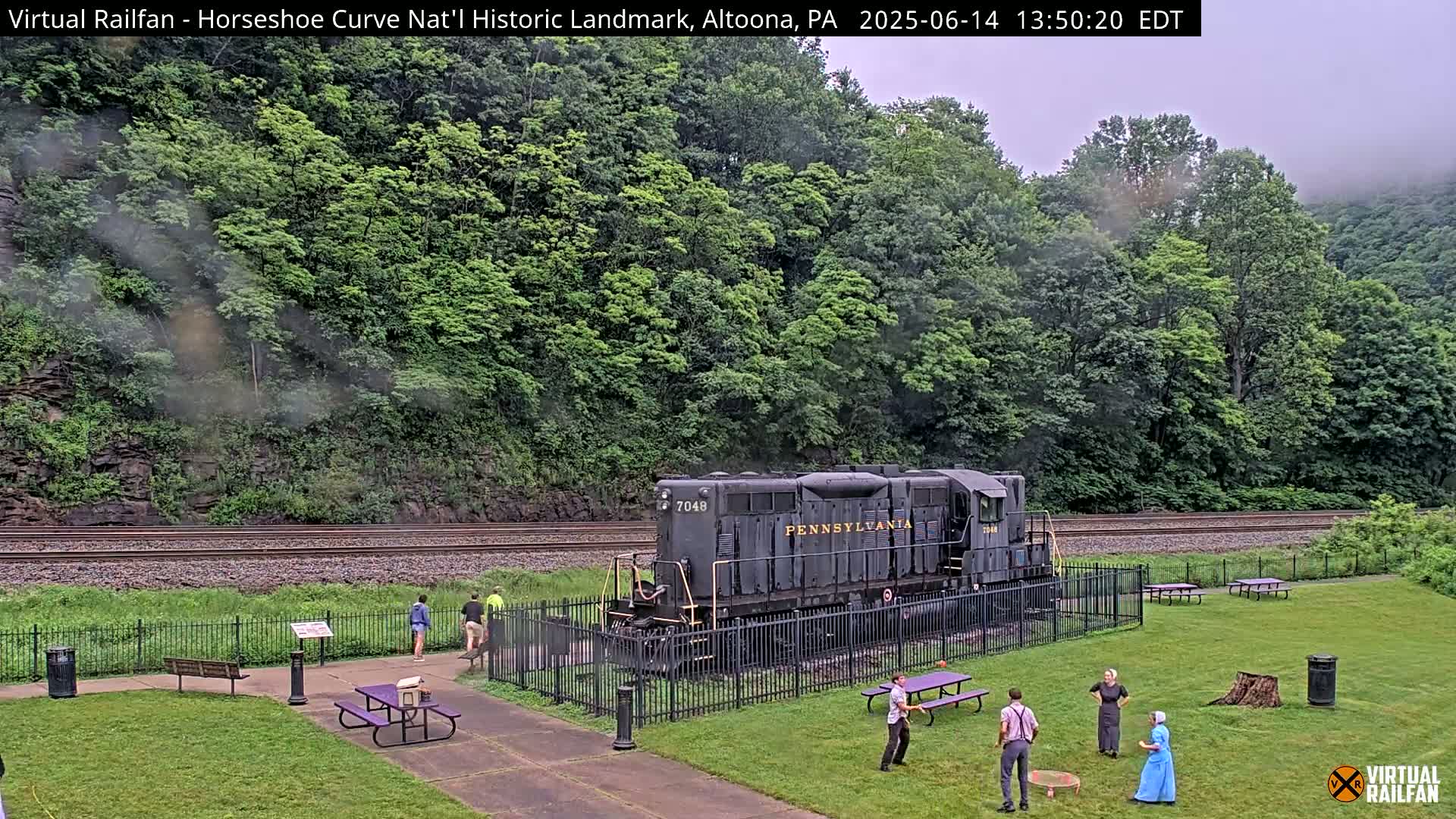 A Pennsylvania Railroad locomotive sits on a grassy area beside train tracks, with several people standing nearby on an overcast day.
