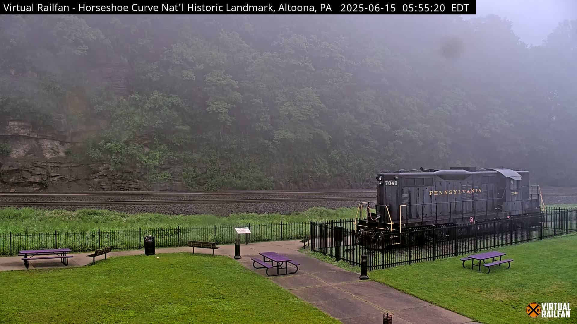 A Pennsylvania Railroad locomotive sits on the tracks behind a grassy park area with picnic tables, all shrouded in a misty fog.