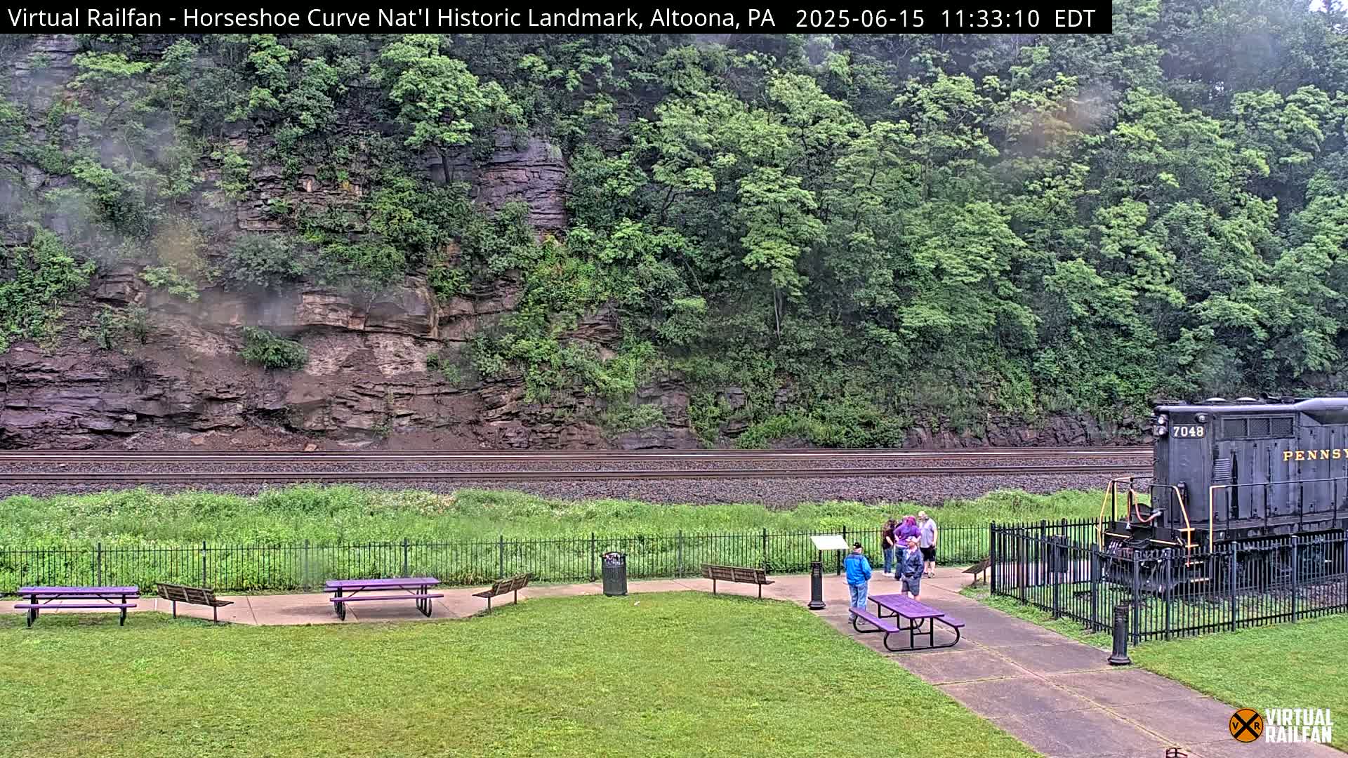 A dark-colored locomotive sits near a grassy area with purple picnic tables and a group of people, all situated before a steep, rocky hillside covered in green foliage, under overcast skies.
