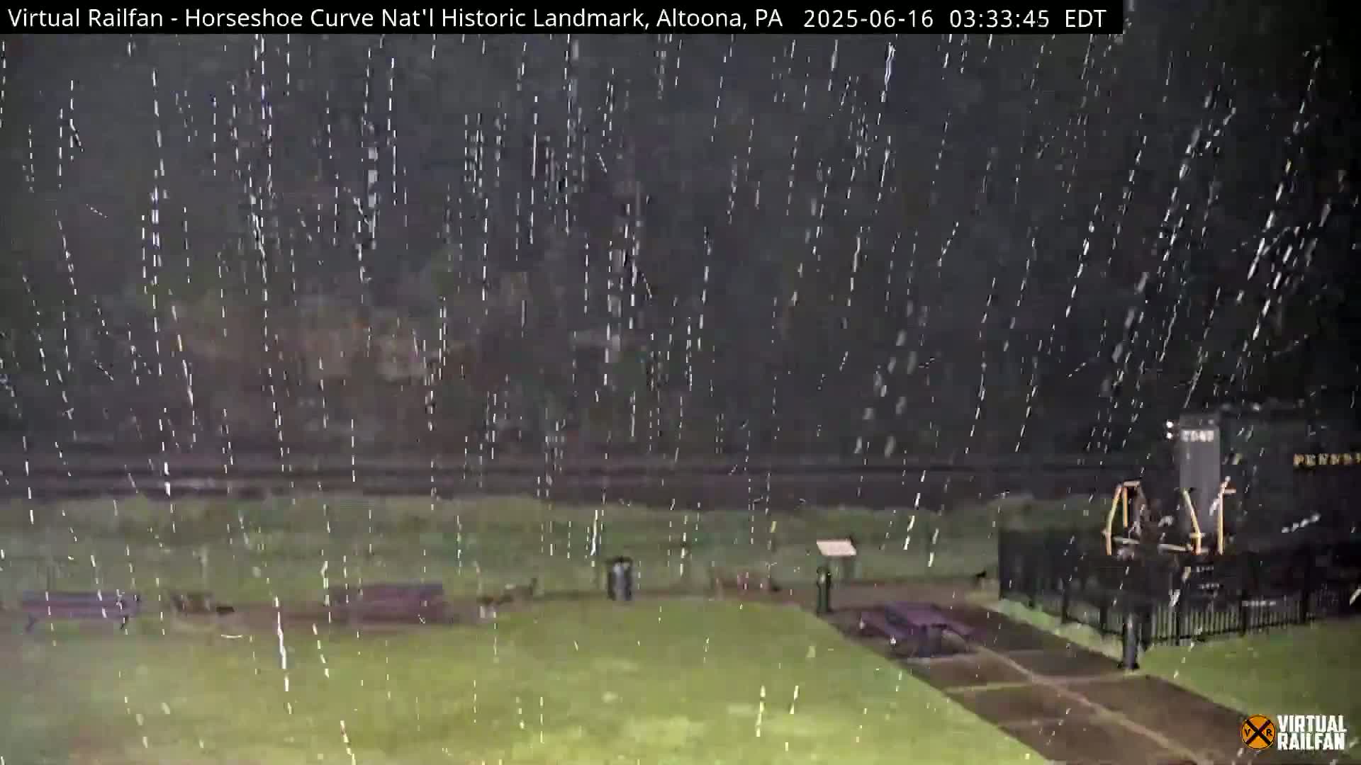 A nighttime scene of a grassy area with park benches and a railroad car visible through heavy rain.