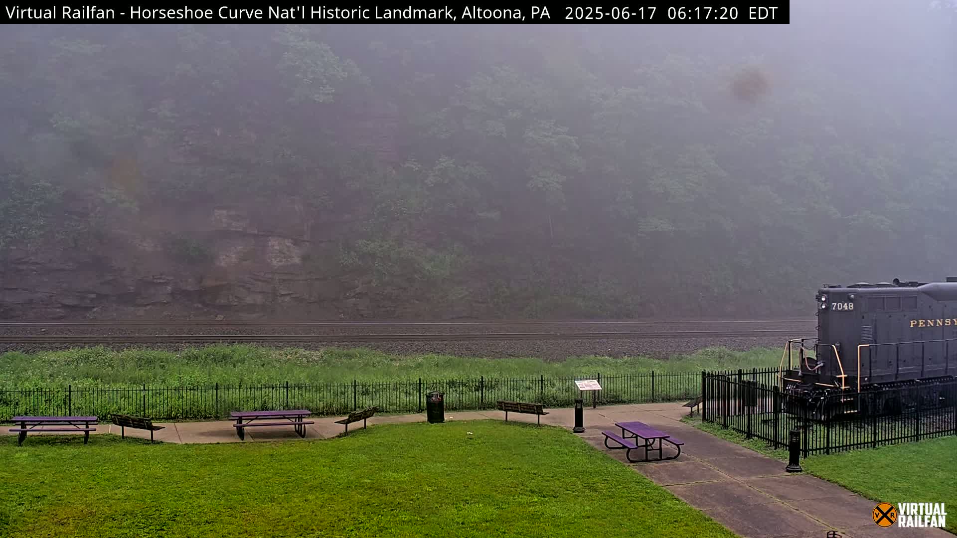 A foggy view of a grassy area with picnic tables, a black metal fence, and a train at the edge of a railroad track, all set against a backdrop of a rocky hillside shrouded in mist.