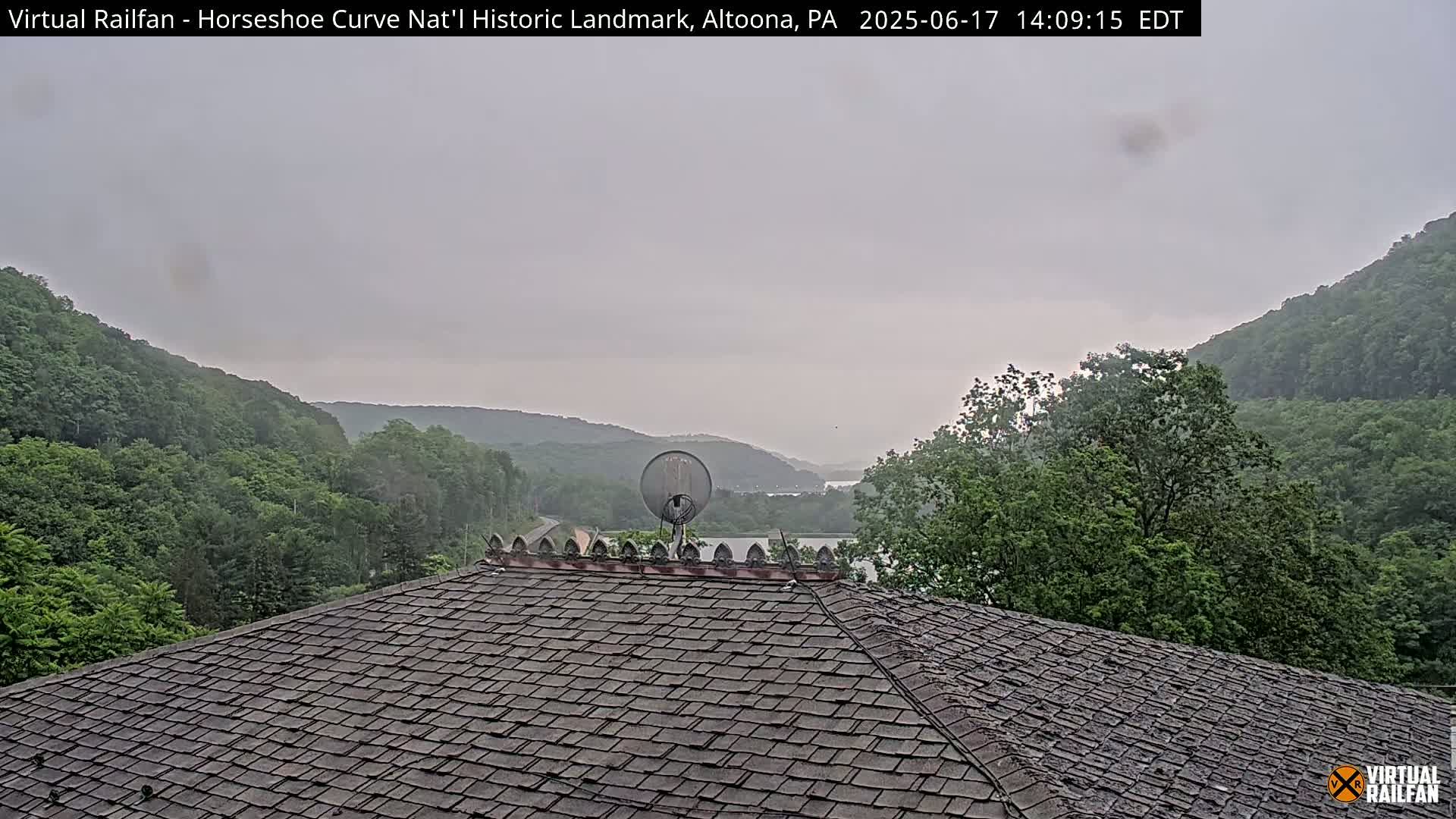 A satellite dish sits atop a shingled roof overlooking a valley of green trees and a body of water under an overcast sky.
