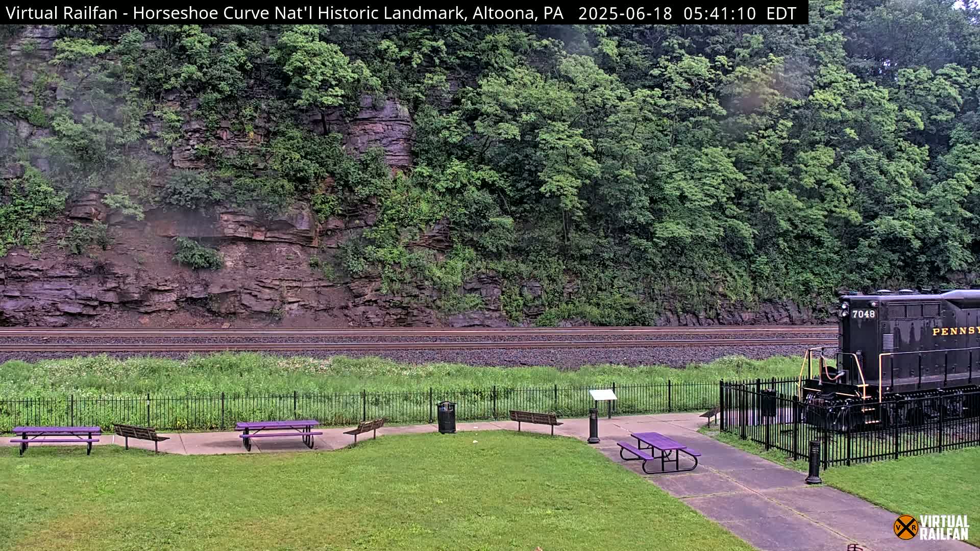 A black locomotive sits near a grassy area with purple picnic tables, beside railroad tracks curving along a rocky hillside covered in lush green vegetation, under overcast skies.