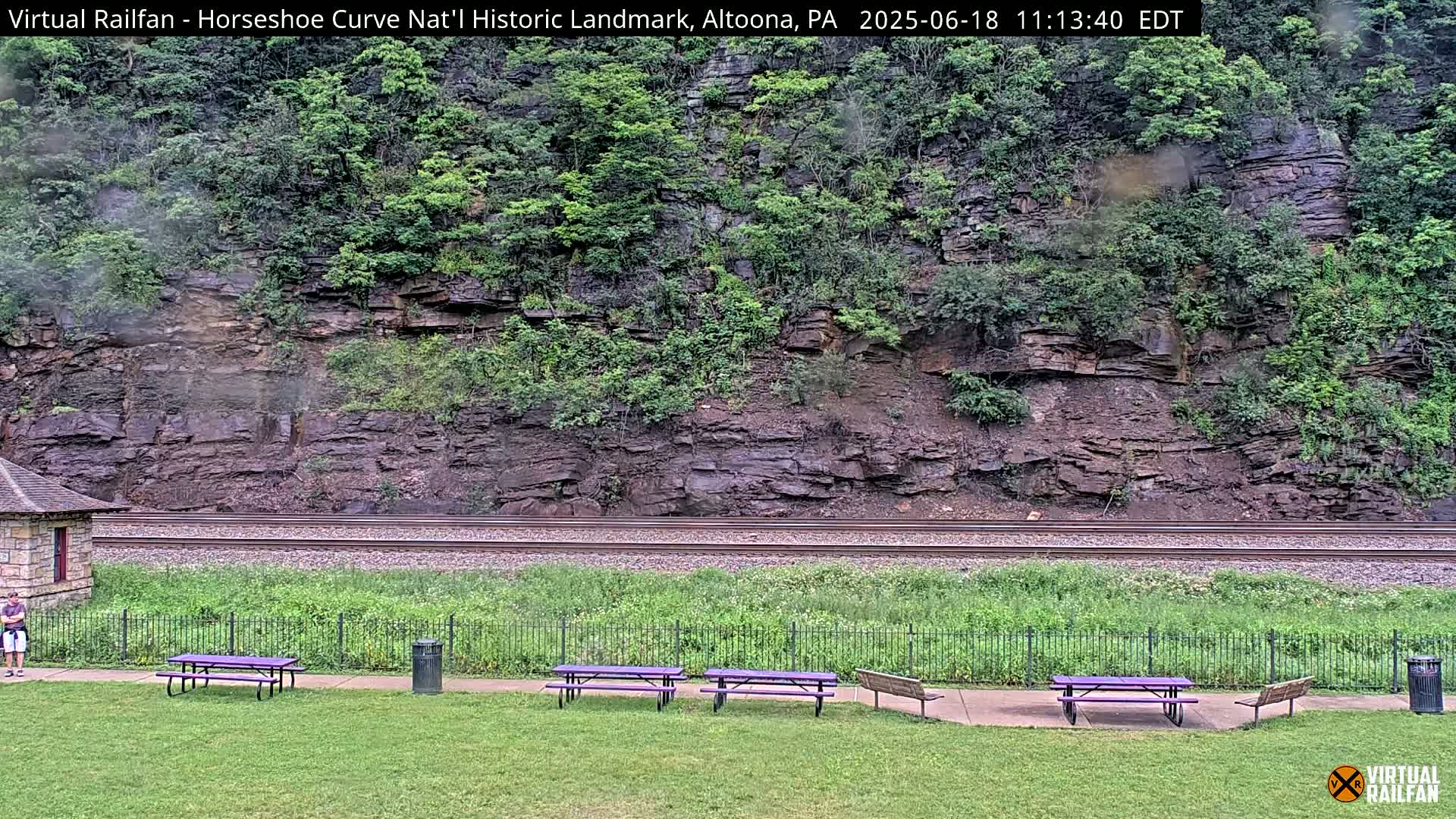 A grassy area with purple picnic tables and a black trash can sits beside railroad tracks that run in front of a rocky hillside covered in green vegetation, under overcast skies.