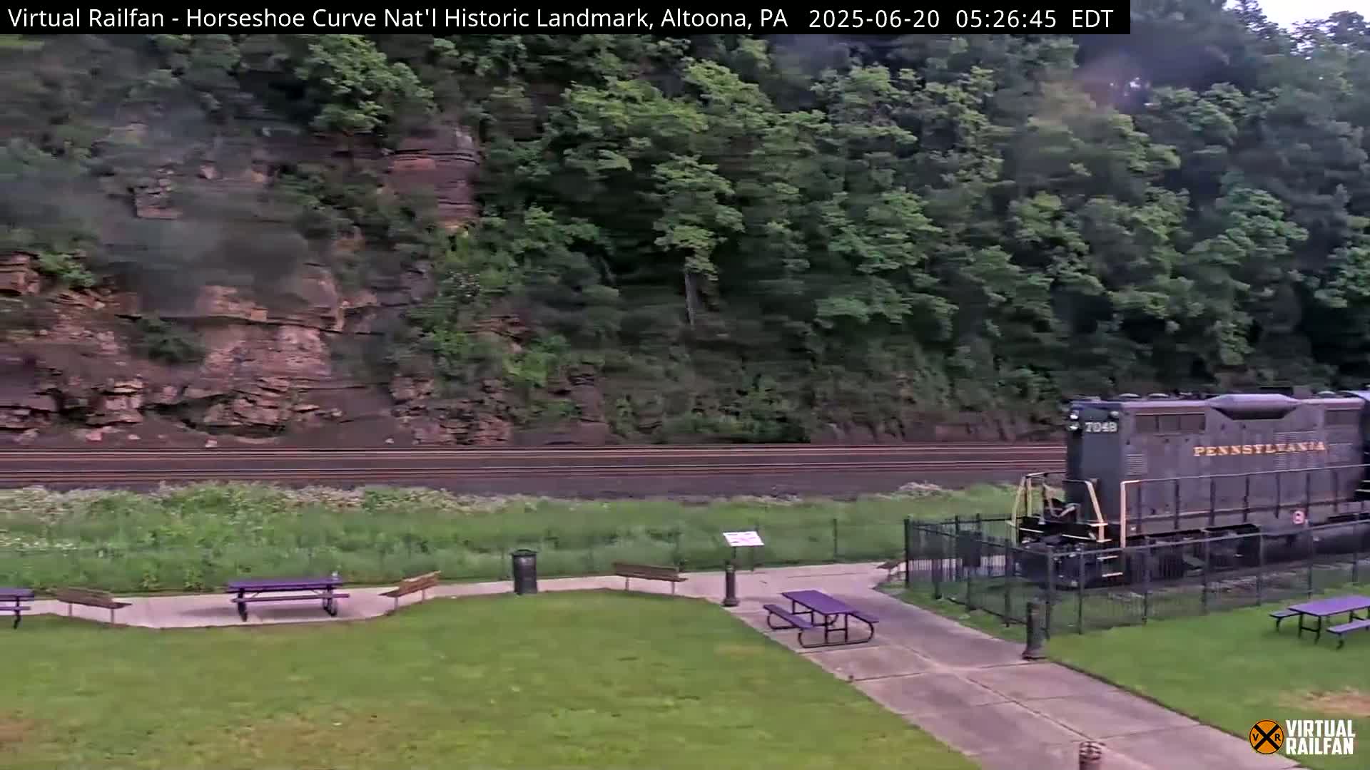 A Pennsylvania Railroad locomotive sits near picnic tables and a paved path beside a grassy area, with a steep, rocky hillside covered in lush green trees in the background.