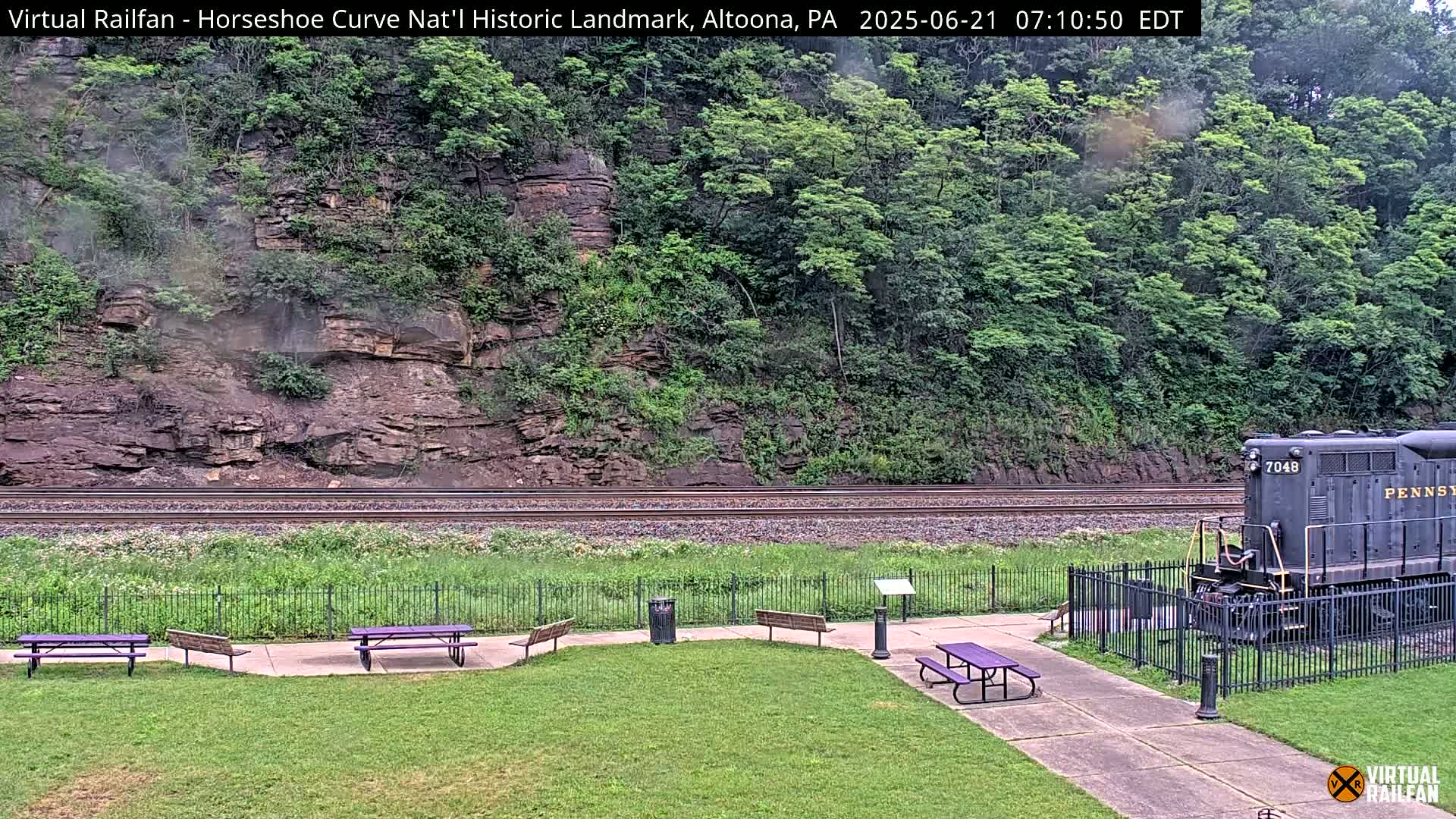 A train sits near a grassy area with picnic tables beside a fenced-in railroad track against a backdrop of a rocky hillside covered in lush green trees, in overcast conditions.