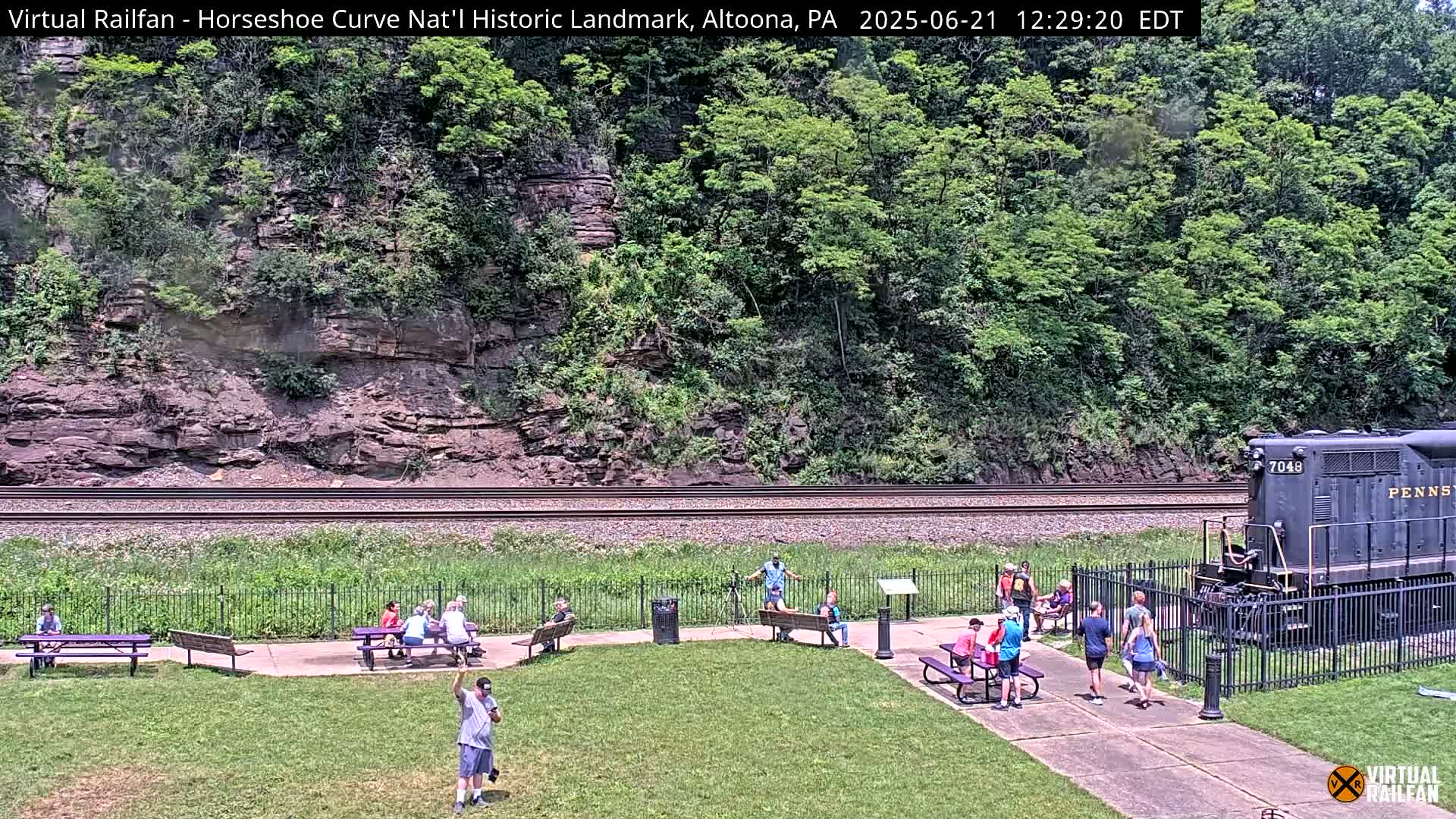 A grassy area with picnic tables and people, situated beside a railroad track with a train visible in the distance, overlooks a steep, rocky hillside covered in lush green trees under a sunny sky.