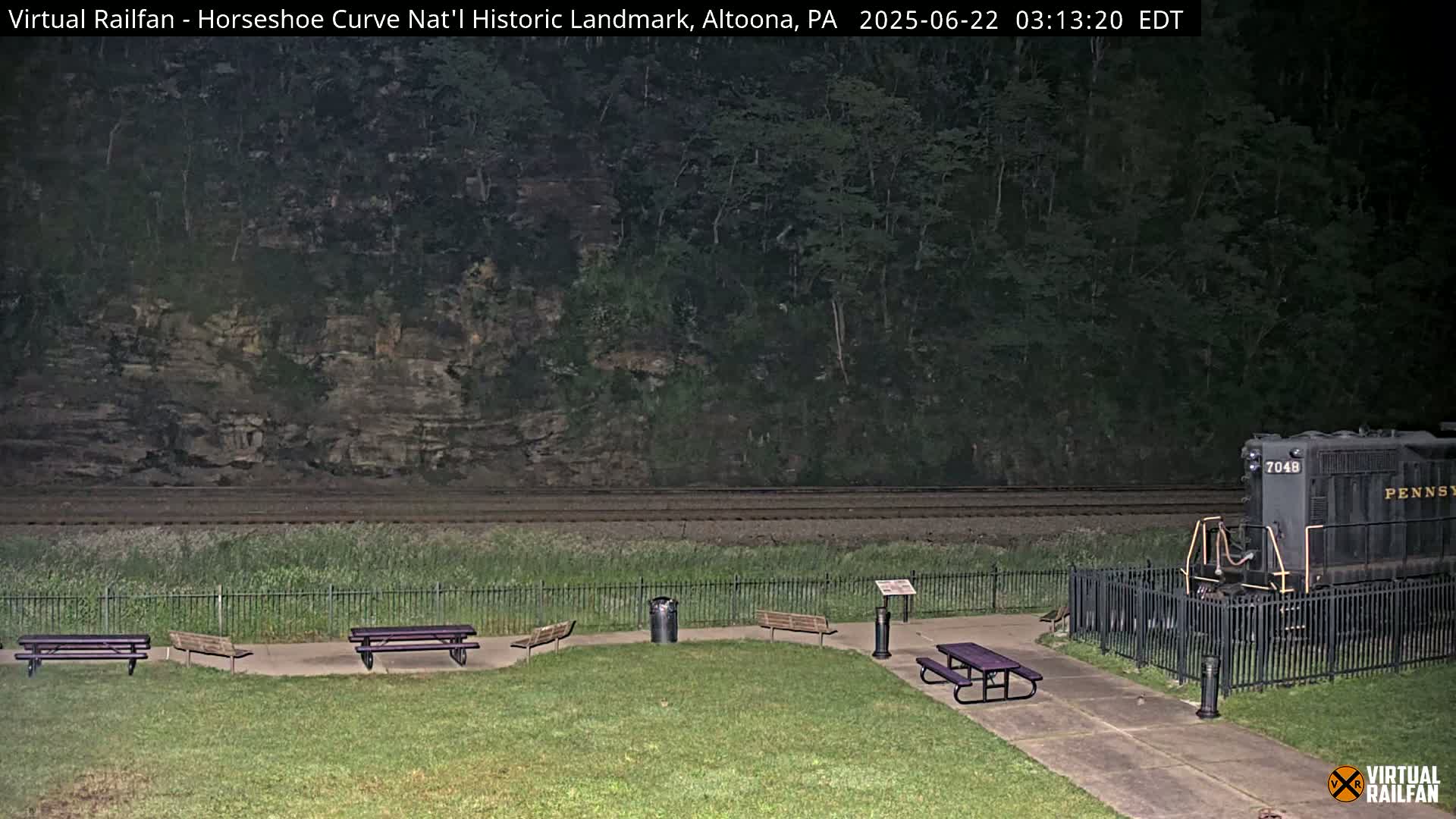 A nighttime view of a grassy area with picnic tables and a trash can, overlooking a railroad track with a locomotive visible beyond a fence, set against a backdrop of a rocky hillside and trees.