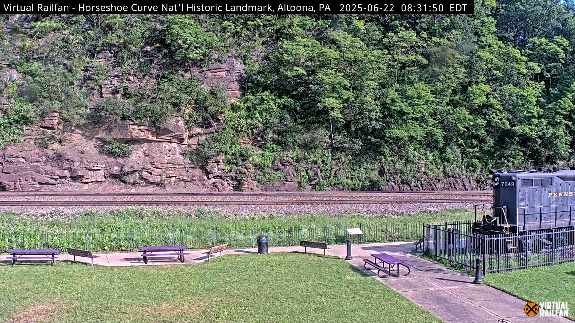 A dark-colored locomotive sits near a grassy area with picnic tables, beside railroad tracks that run along a rocky hillside covered in lush green vegetation, under a sunny sky.