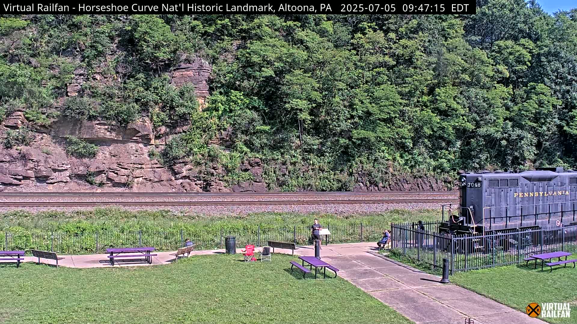 A Pennsylvania Railroad locomotive sits near a grassy area with picnic tables and a few people, all situated beside a railway track running alongside a rocky hillside with lush green vegetation under a sunny sky.