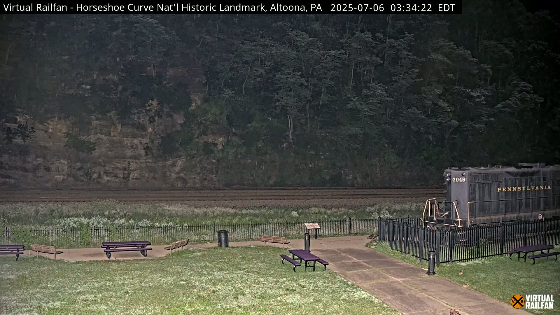 A dark-colored locomotive sits behind a black fence next to a grassy area with several picnic tables, all set against a backdrop of a wooded hillside under a nighttime sky.