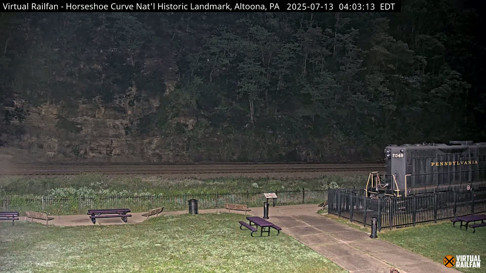 A dark-colored locomotive sits behind a fence in a grassy area with picnic tables, near railroad tracks and a wooded hillside at night.