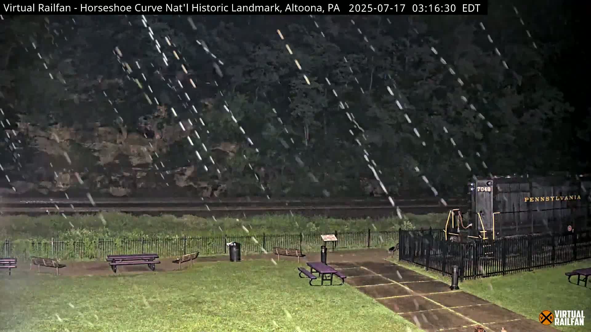 A nighttime view of a park with picnic tables, a fenced-in train, and heavy rain falling.
