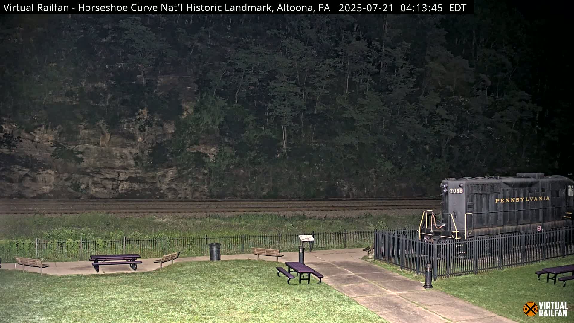A dark-colored diesel locomotive sits behind a black fence near picnic tables on a grassy area beside a railroad track at night.
