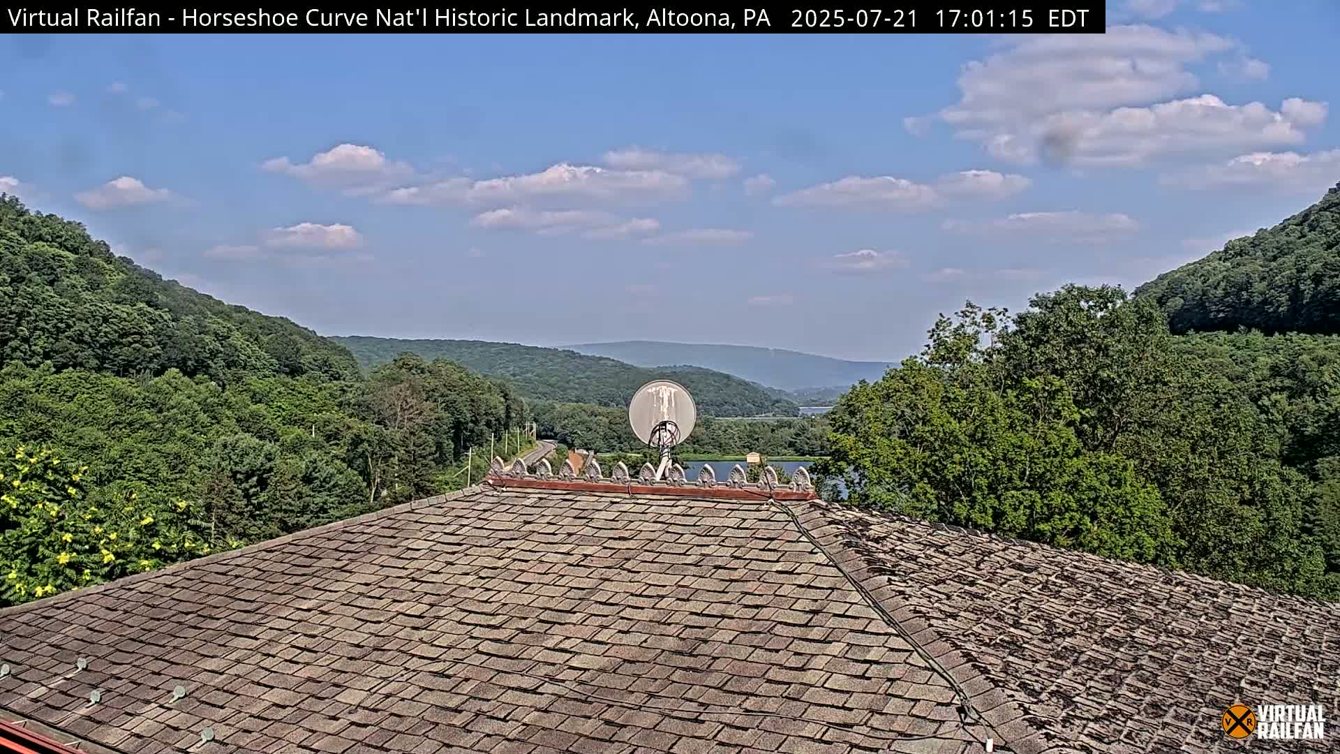 A shingled roof overlooks a partly cloudy sky, green hills, a road, and a body of water.