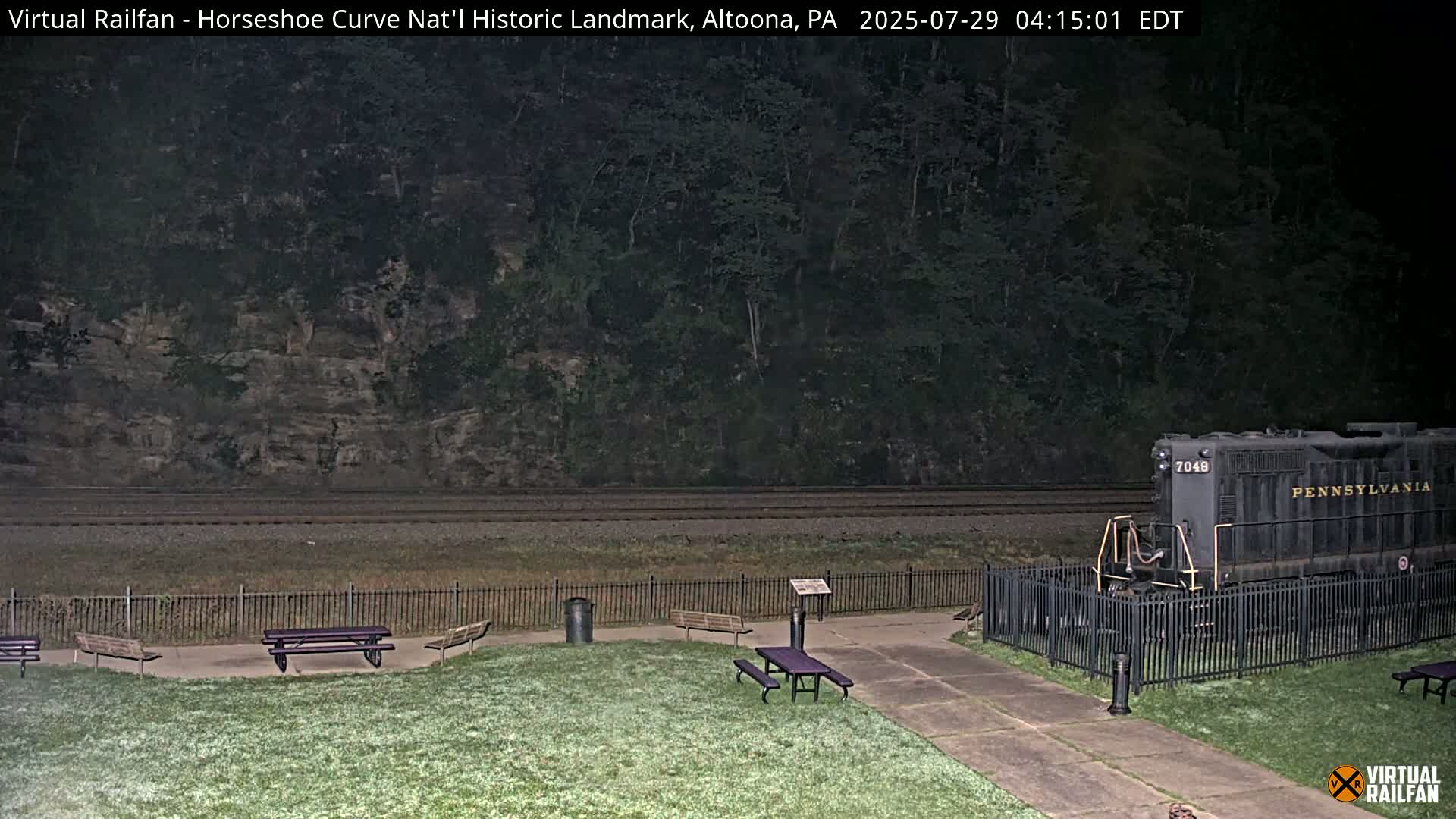 A dark-colored locomotive sits behind a black metal fence in a grassy area with several picnic tables, all under a nighttime sky.