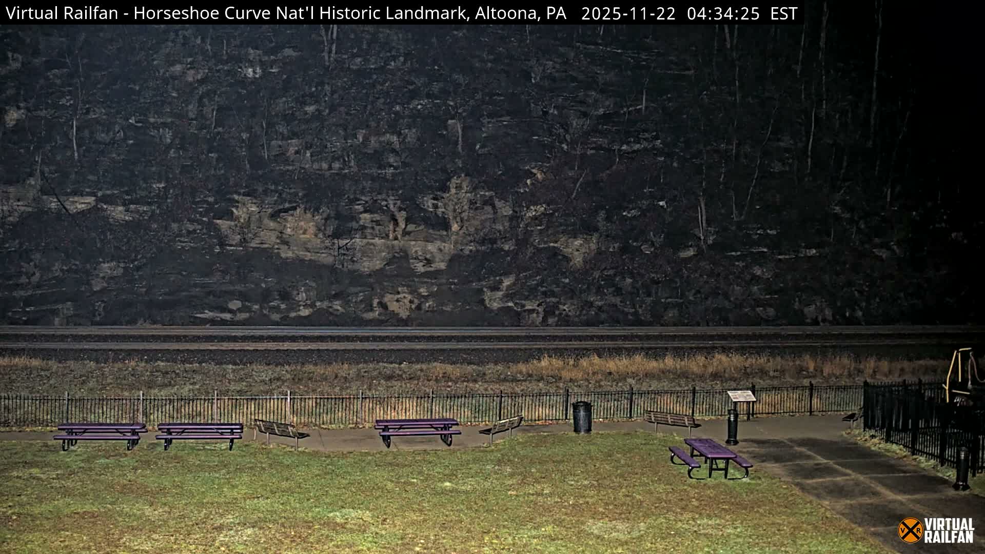 The image displays a damp park at night featuring purple picnic tables and benches in the foreground, three parallel railroad tracks traversing the midground, and a dark, rocky, tree-covered hillside rising in the background under a clear sky.
