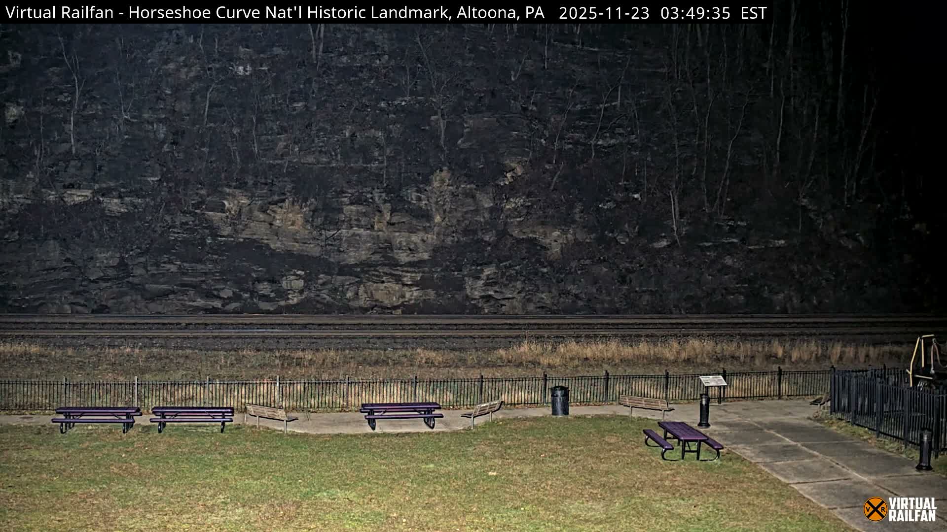 At night under an overcast sky, three railroad tracks run along the base of a steep, bare forested hillside behind a black metal fence, which separates the tracks from a grassy area featuring purple picnic tables, wooden benches, and a concrete pathway.